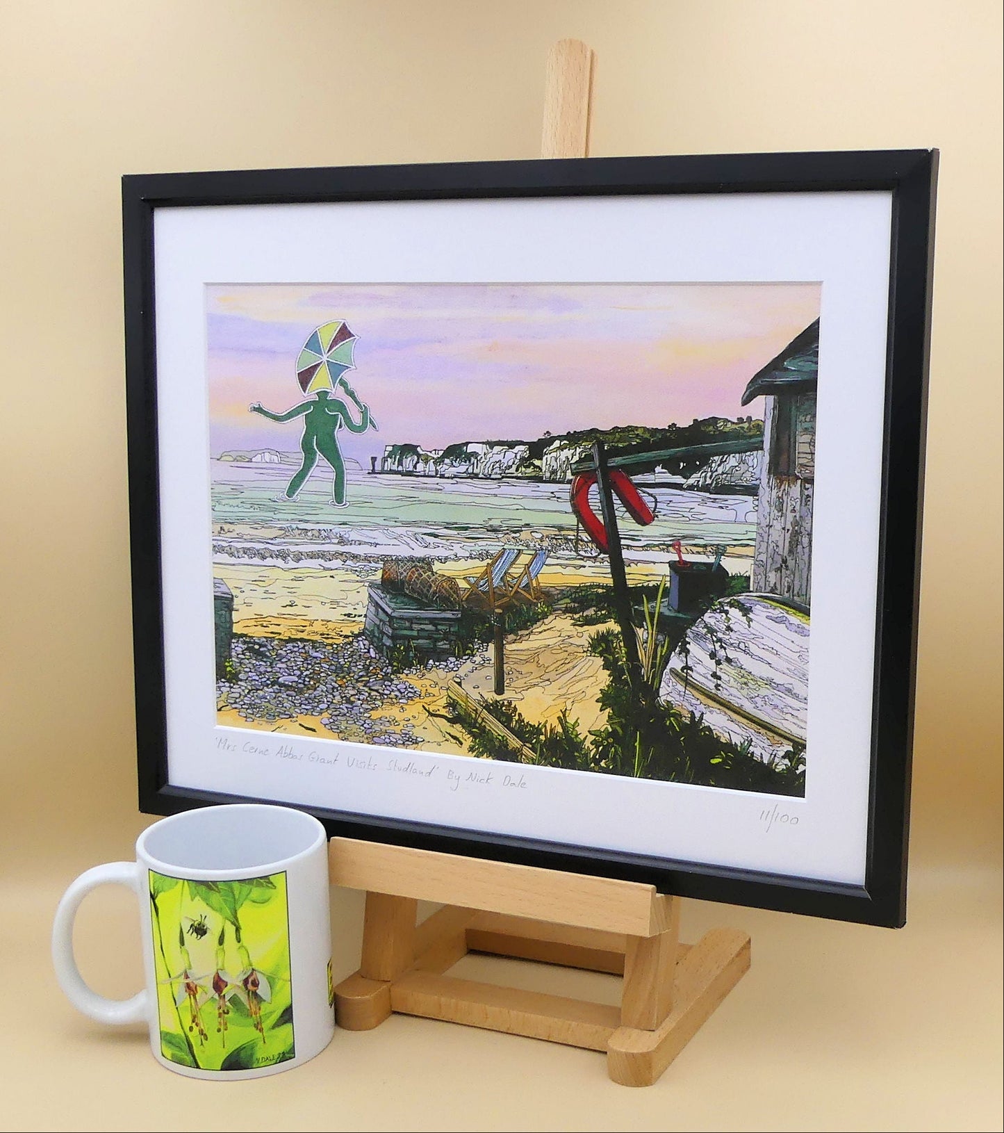 Image of a Medium Art Print featuring a copy of the painting 'Mrs Cerne Abbas Giant Visits Studland' by Nick Dale. The print imagines the Cerne Abbas Giant's wife visiting the seaside at Studland Beach; with Old Harry Rocks and the Isle of Wight in the distance. The print is situated on a small easel in a black frame next to a mug.