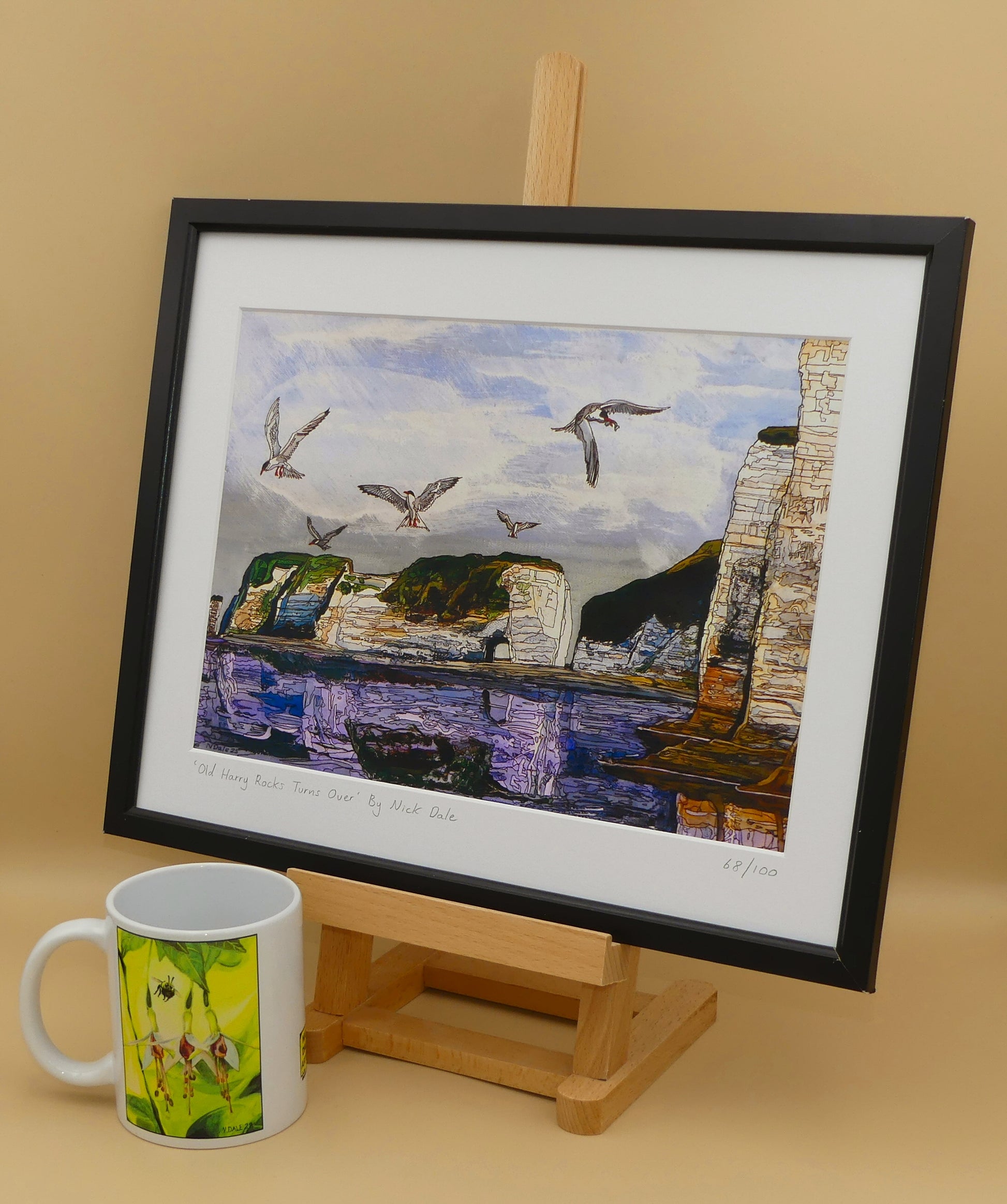 Image of a Medium Art Print featuring a copy of the painting 'Old Harry Rocks Turns Over' by Nick Dale. The print depicts the view on a walk in March 2025 when the tide was completely out so I could walk to Old Harry Rocks. I never realised that that was ever possible. I had to add in the water when I painted it afterwards as it was completely missing. Turns also added to taste. The print is situated on a small easel in a black frame next to a mug.