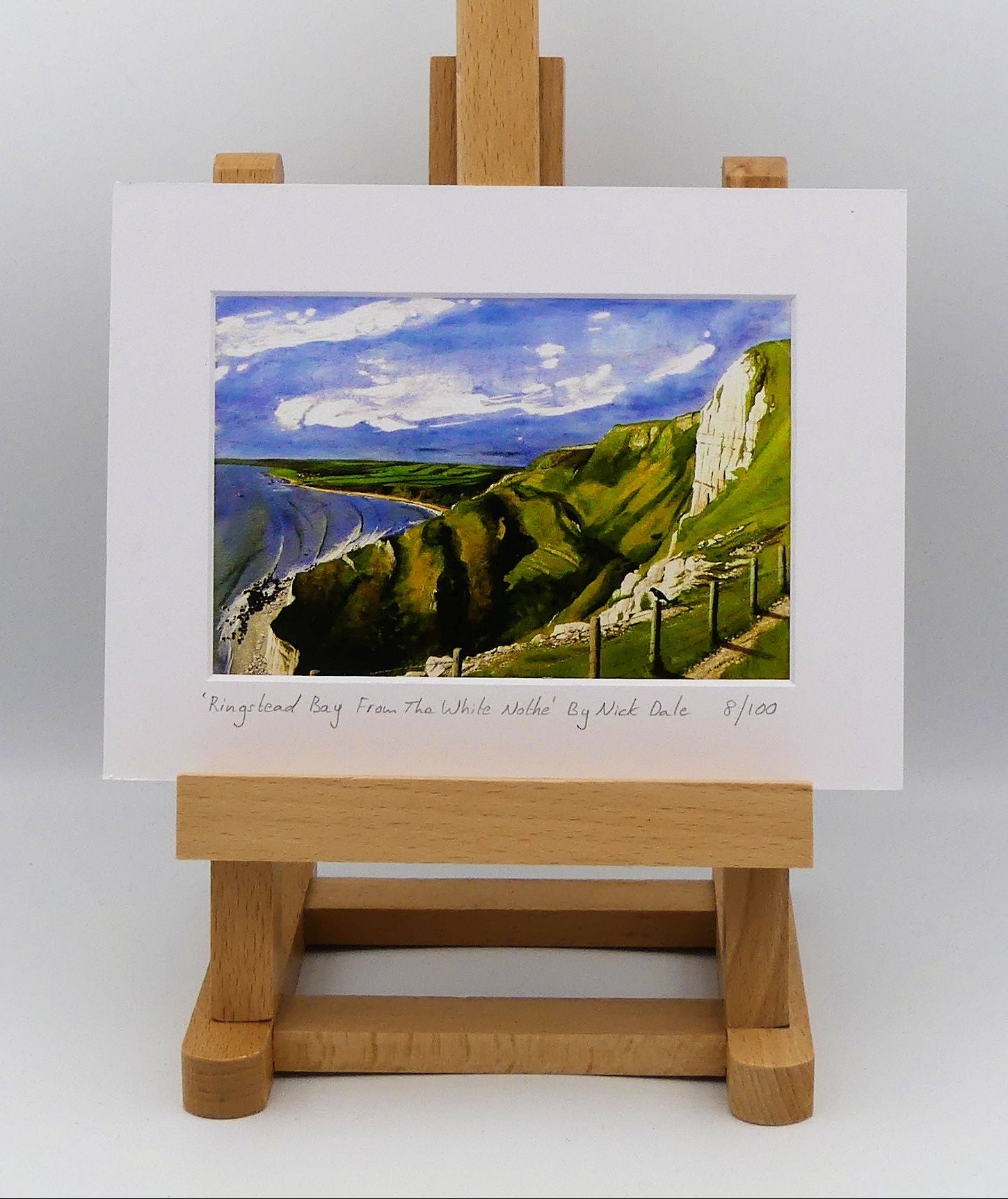 Image of a Small Art Print featuring a copy of the painting 'Ringstead Bay From The White Nothe' by Nick Dale. The print depicts a view along the Jurassic Coast from the White Nothe chalk headland east of Ringstead Bay. The view looks west over the bay to the village of Ringstead & beyond.  The print is situated on a small easel.