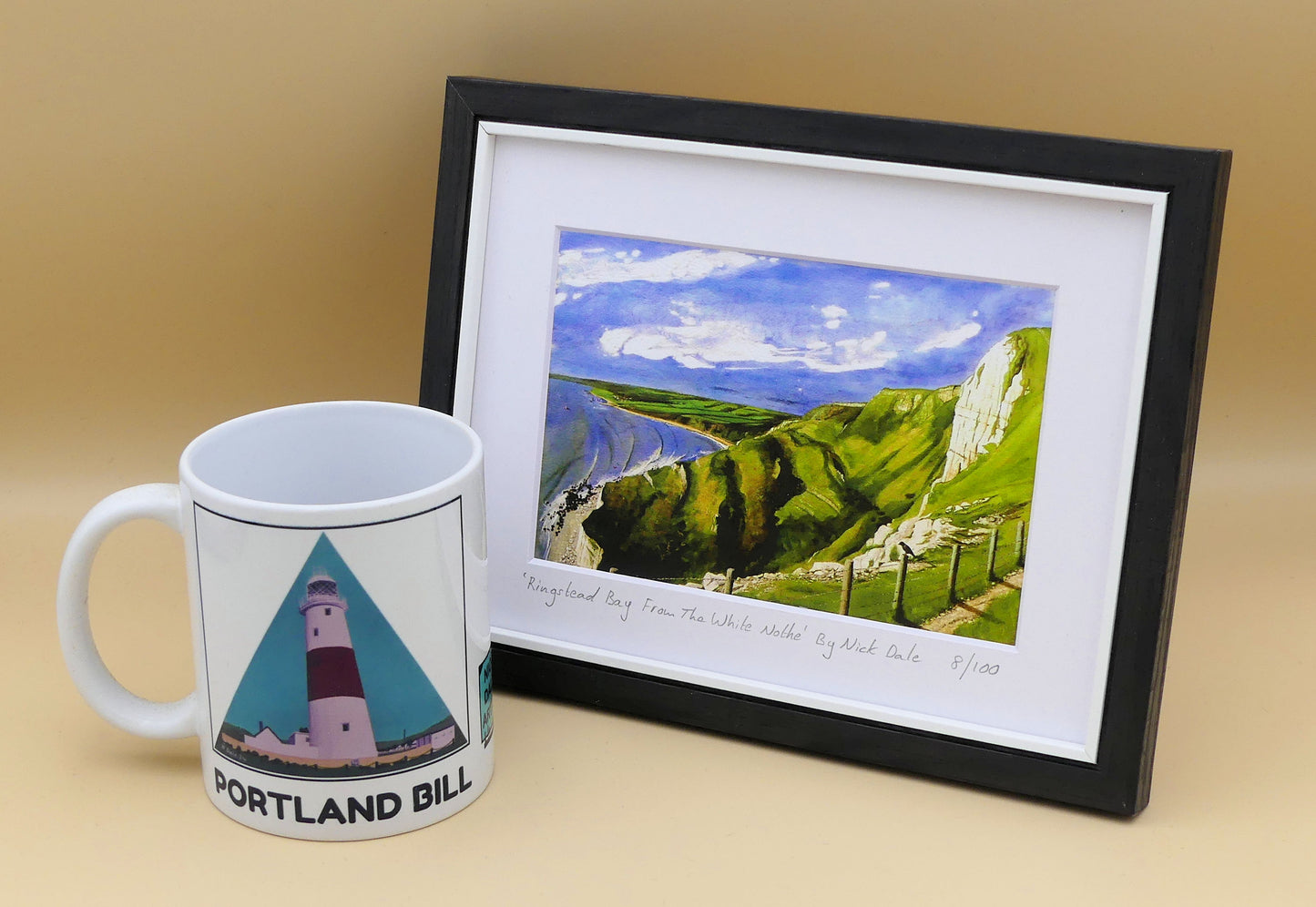 Image of a Small Art Print featuring a copy of the painting 'Ringstead Bay From The White Nothe' by Nick Dale. The print depicts a view along the Jurassic Coast from the White Nothe chalk headland east of Ringstead Bay. The view looks west over the bay to the village of Ringstead & beyond. The print is situated in a black frame next to a mug.