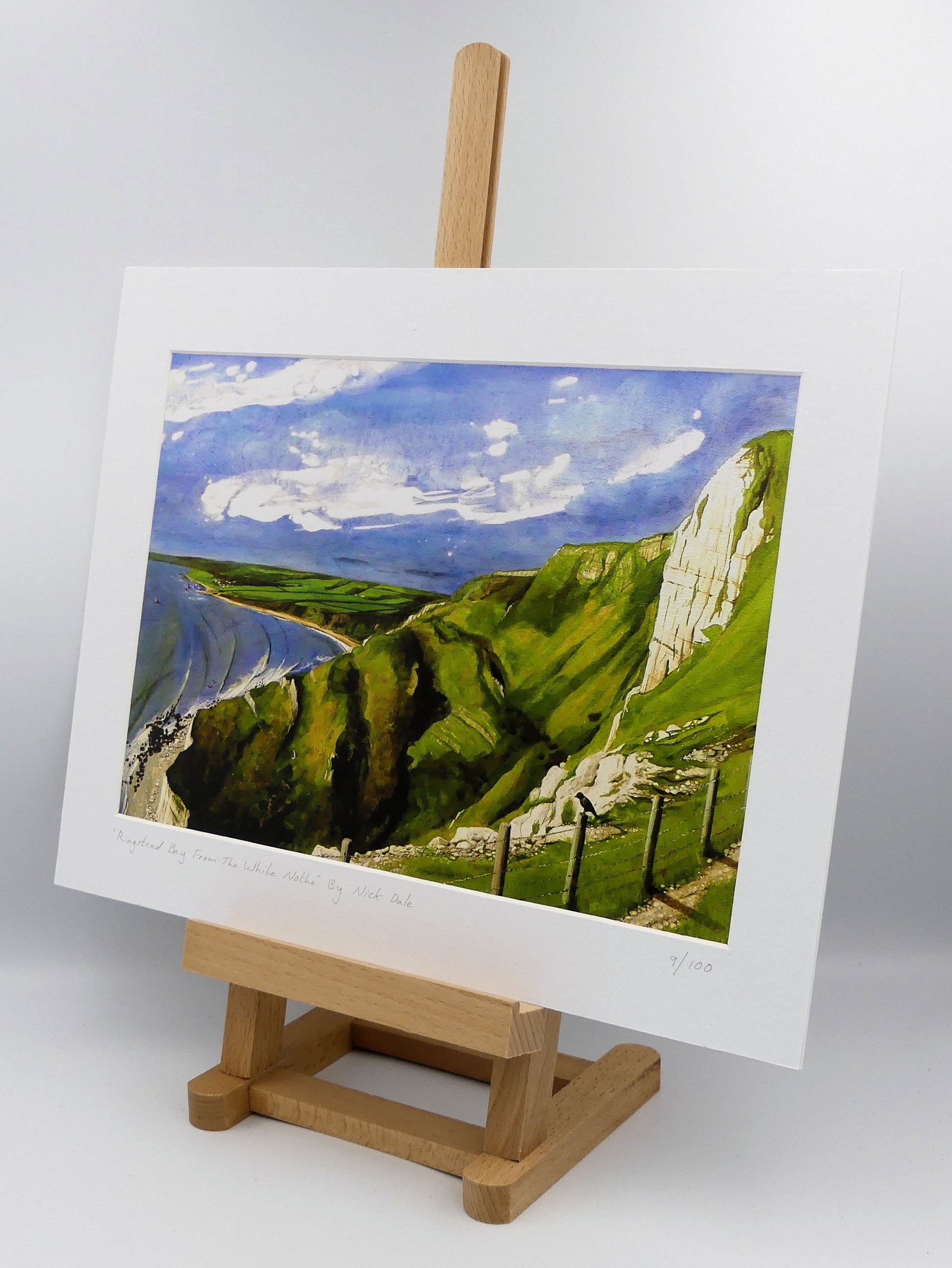 Image of a Medium Art Print featuring a copy of the painting 'Ringstead Bay From The White Nothe' by Nick Dale. The print depicts a view along the Jurassic Coast from the White Nothe chalk headland east of Ringstead Bay. The view looks west over the bay to the village of Ringstead & beyond. The print is situated on a small easel.