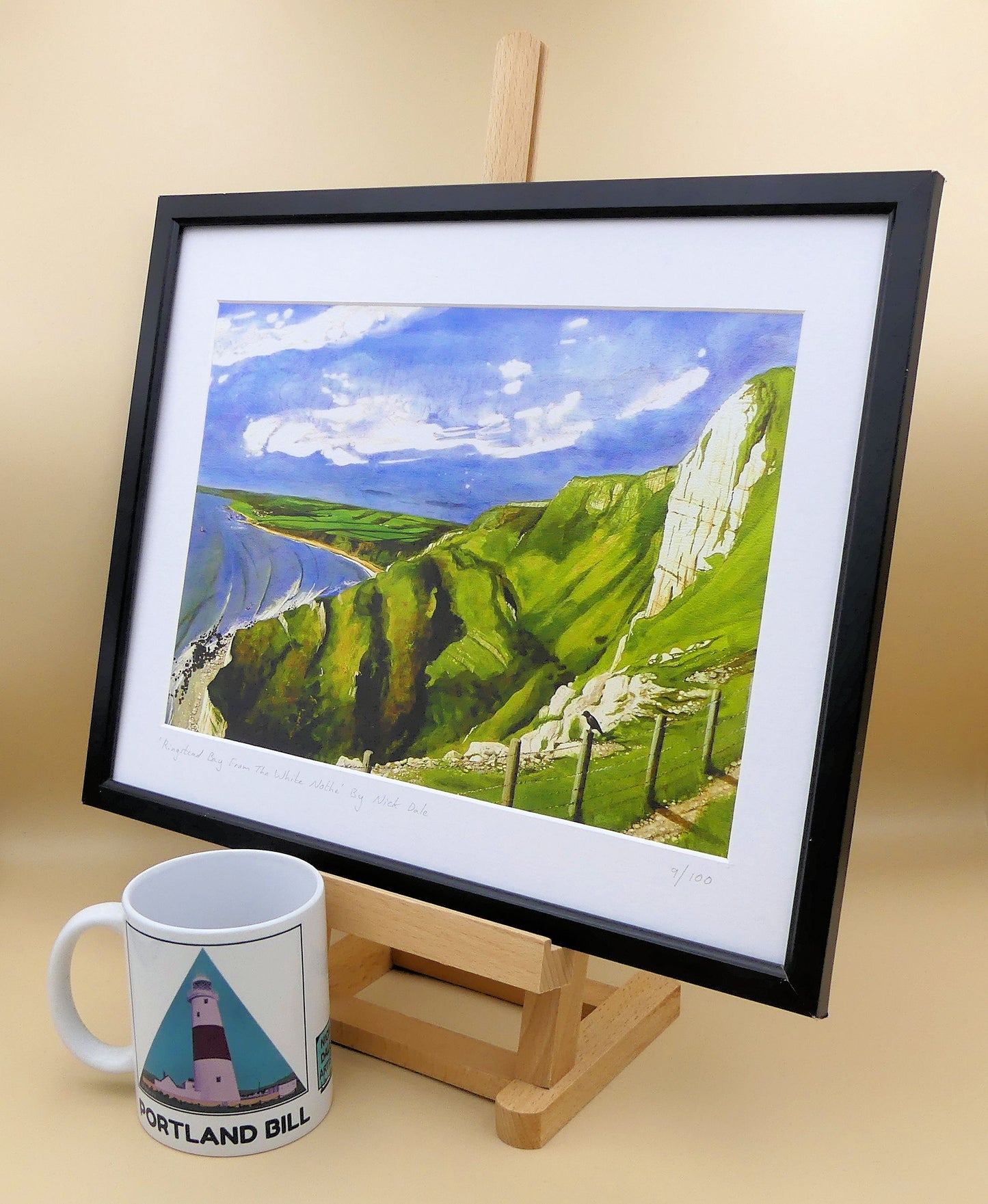 Image of a Medium Art Print featuring a copy of the painting 'Ringstead Bay From The White Nothe' by Nick Dale. The print depicts a view along the Jurassic Coast from the White Nothe chalk headland east of Ringstead Bay. The view looks west over the bay to the village of Ringstead & beyond. The print is situated on a small easel in a black frame next to a mug.