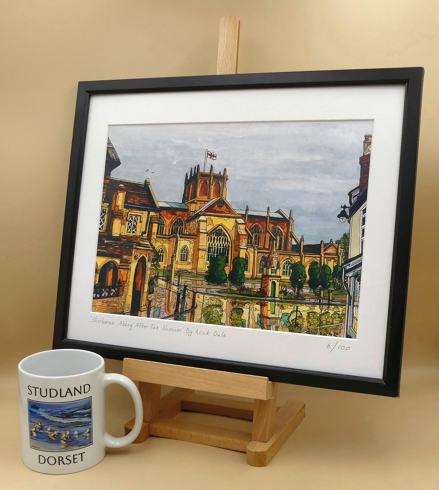 Image of a Medium Art Print featuring a copy of the painting 'Sherborne Abbey After The Shower' by Nick Dale. The print depicts a view of Sherborne Abbey (also known as the Abbey Church of St. Mary the Virgin), in Sherborne, Dorset. On the left hand side can be seen St Johns' Almshouse.
The print is situated on a small easel in a black frame next to a mug.