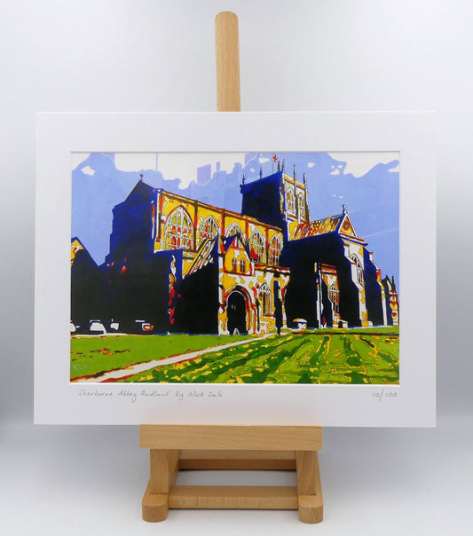 Image of a Medium Art Print featuring a copy of the painting 'Sherborne Abbey Radiant' by Nick Dale. The print depicts Sherborne Abbey in bright primary colours with strong shadows and a brightly lit south-facing aspect. The print is situated on a small easel.