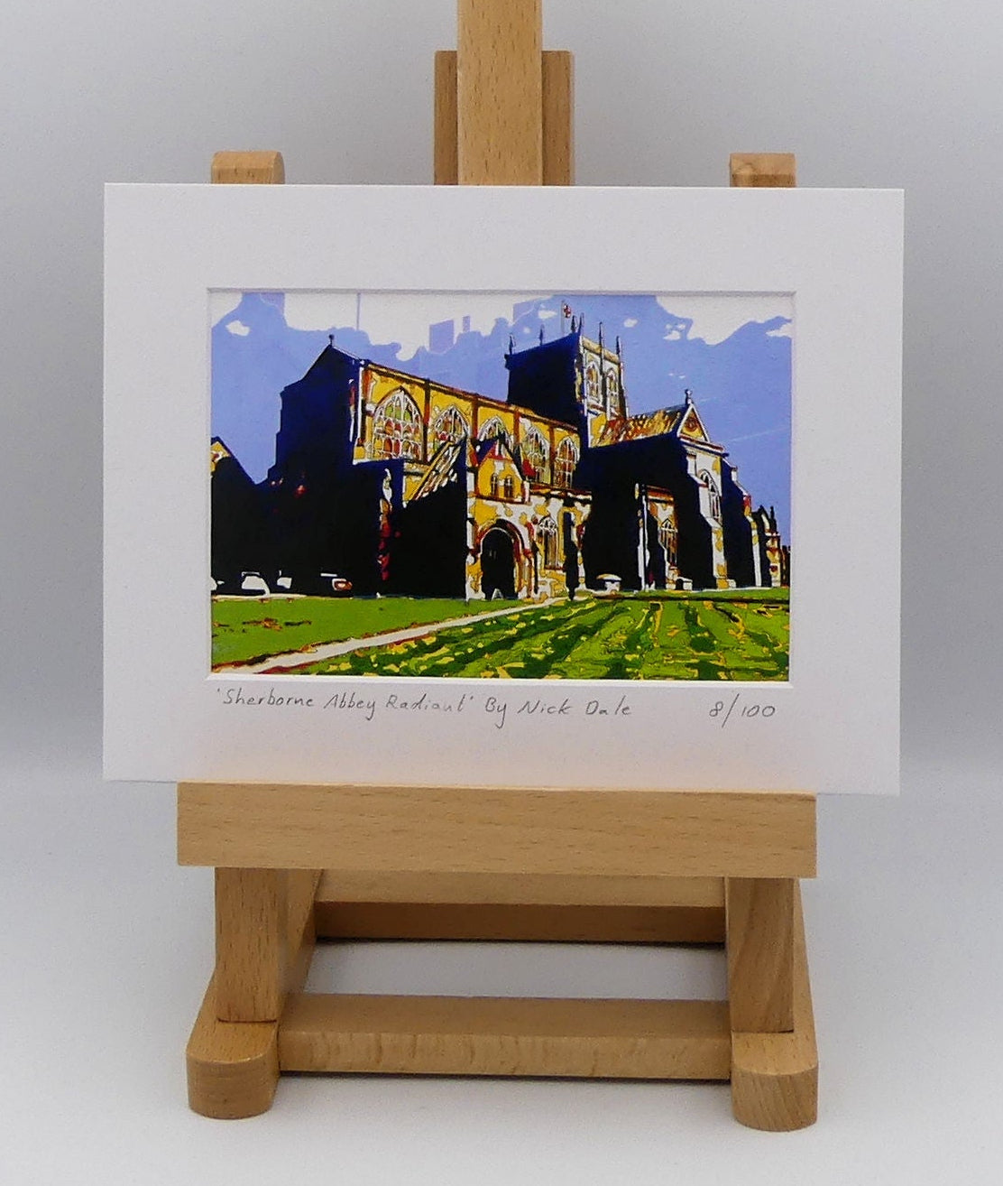 Image of a Small Art Print featuring a copy of the painting 'Sherborne Abbey Radiant' by Nick Dale. The print depicts Sherborne Abbey in bright primary colours with strong shadows and a brightly lit south-facing aspect.  The print is situated on a small easel.