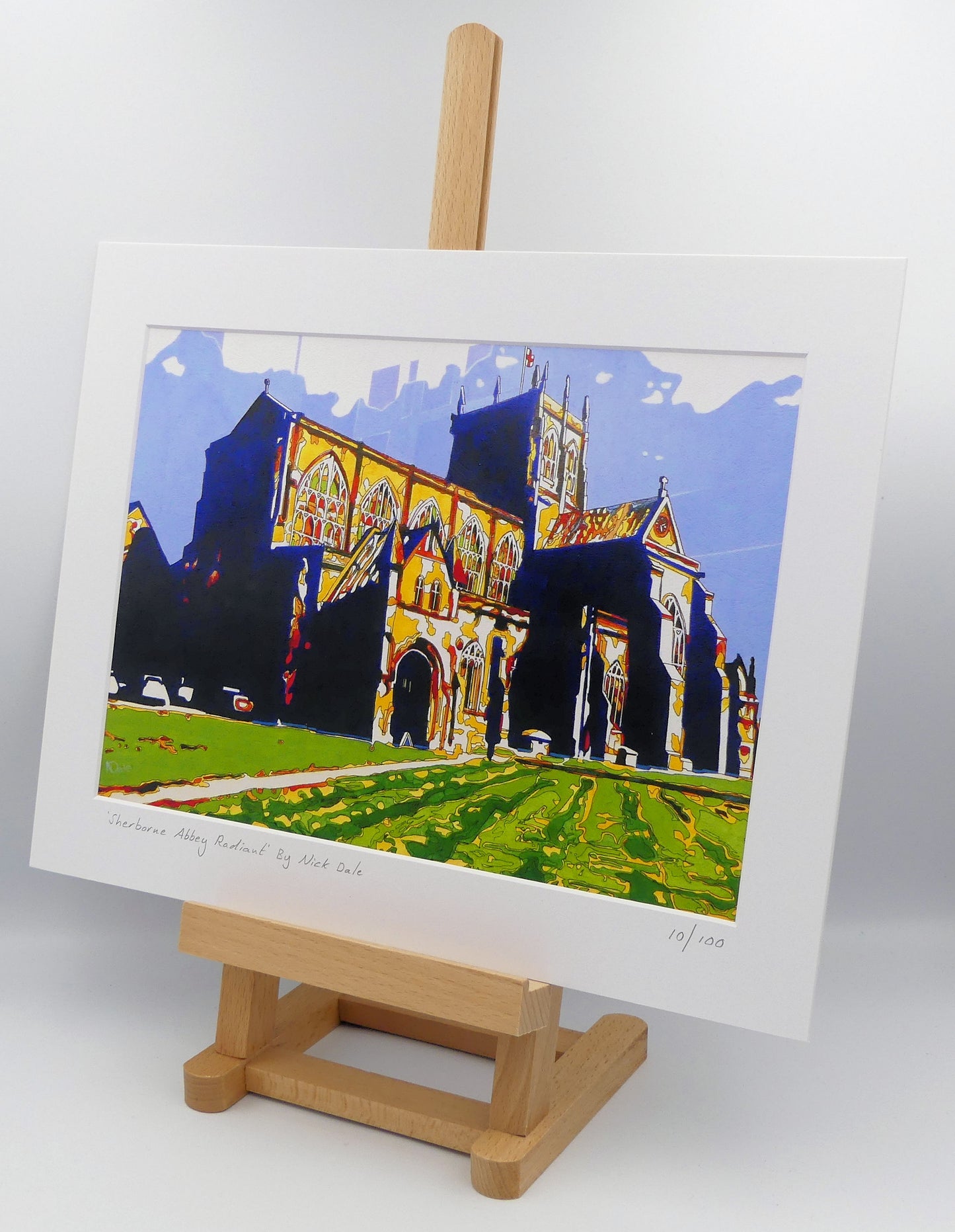 Image of a Medium Art Print featuring a copy of the painting 'Sherborne Abbey Radiant' by Nick Dale. The print depicts Sherborne Abbey in bright primary colours with strong shadows and a brightly lit south-facing aspect. The print is situated on a small easel.