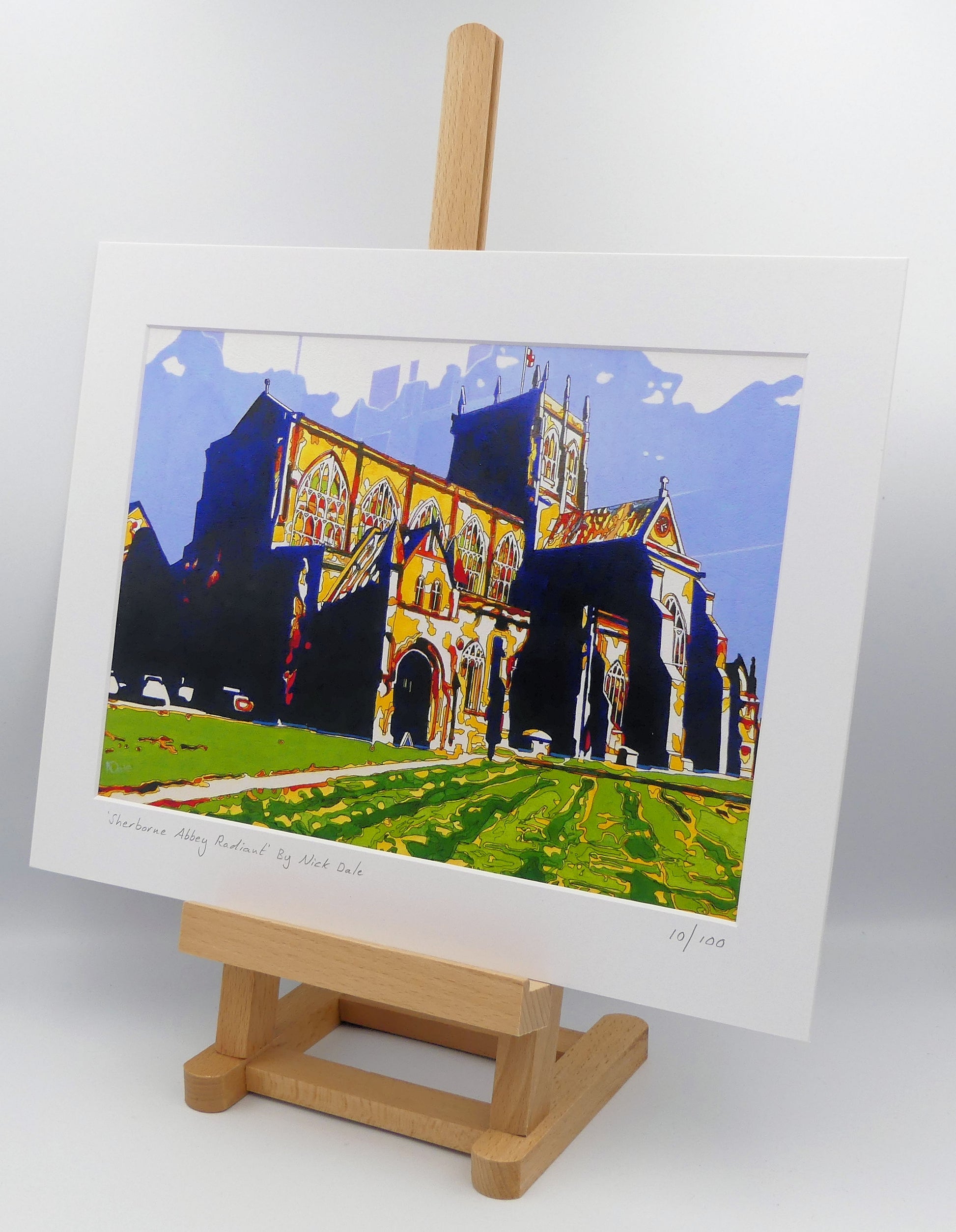 Image of a Medium Art Print featuring a copy of the painting 'Sherborne Abbey Radiant' by Nick Dale. The print depicts Sherborne Abbey in bright primary colours with strong shadows and a brightly lit south-facing aspect. The print is situated on a small easel.