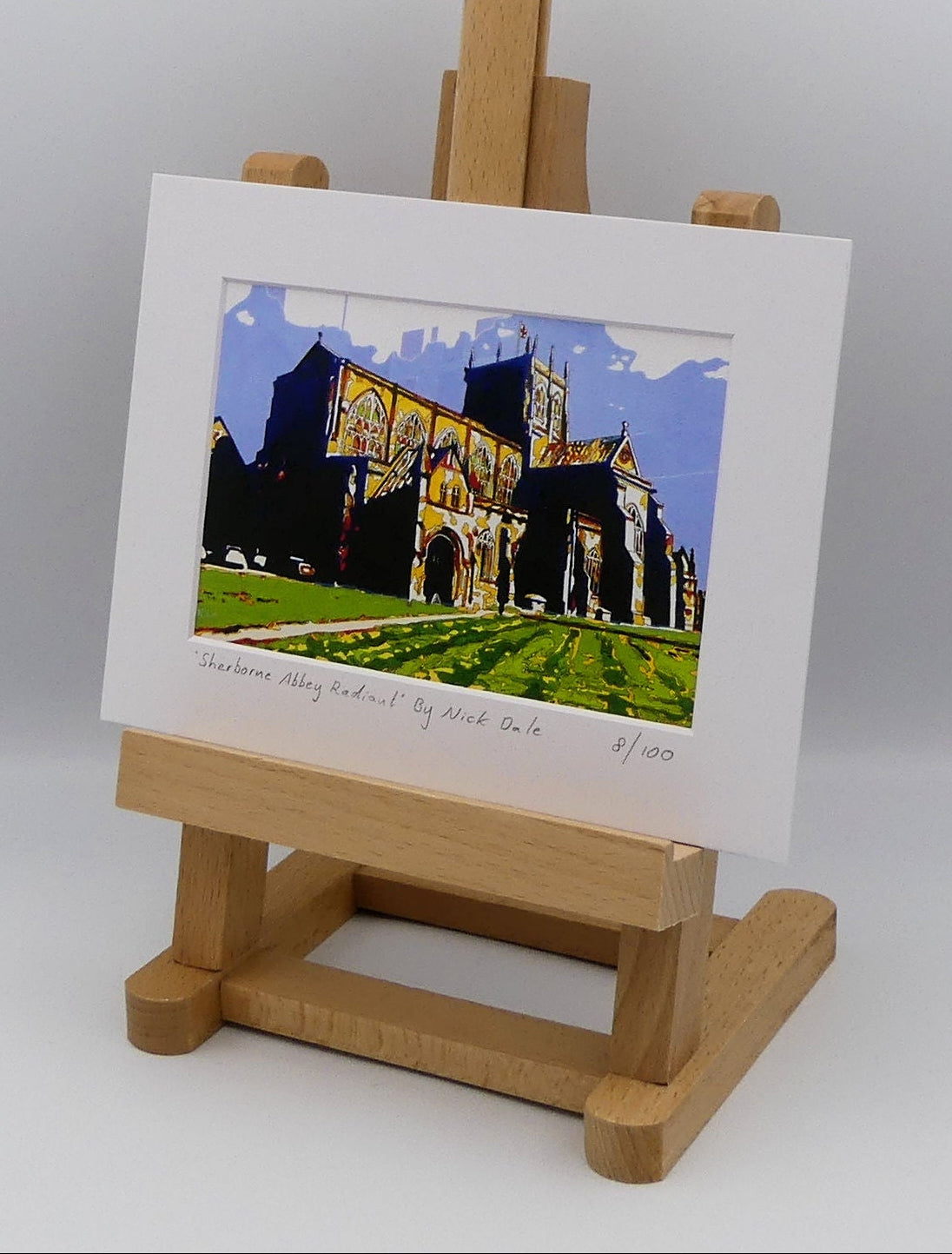 Image of a Small Art Print featuring a copy of the painting 'Sherborne Abbey Radiant' by Nick Dale. The print depicts Sherborne Abbey in bright primary colours with strong shadows and a brightly lit south-facing aspect. The print is situated on a small easel.