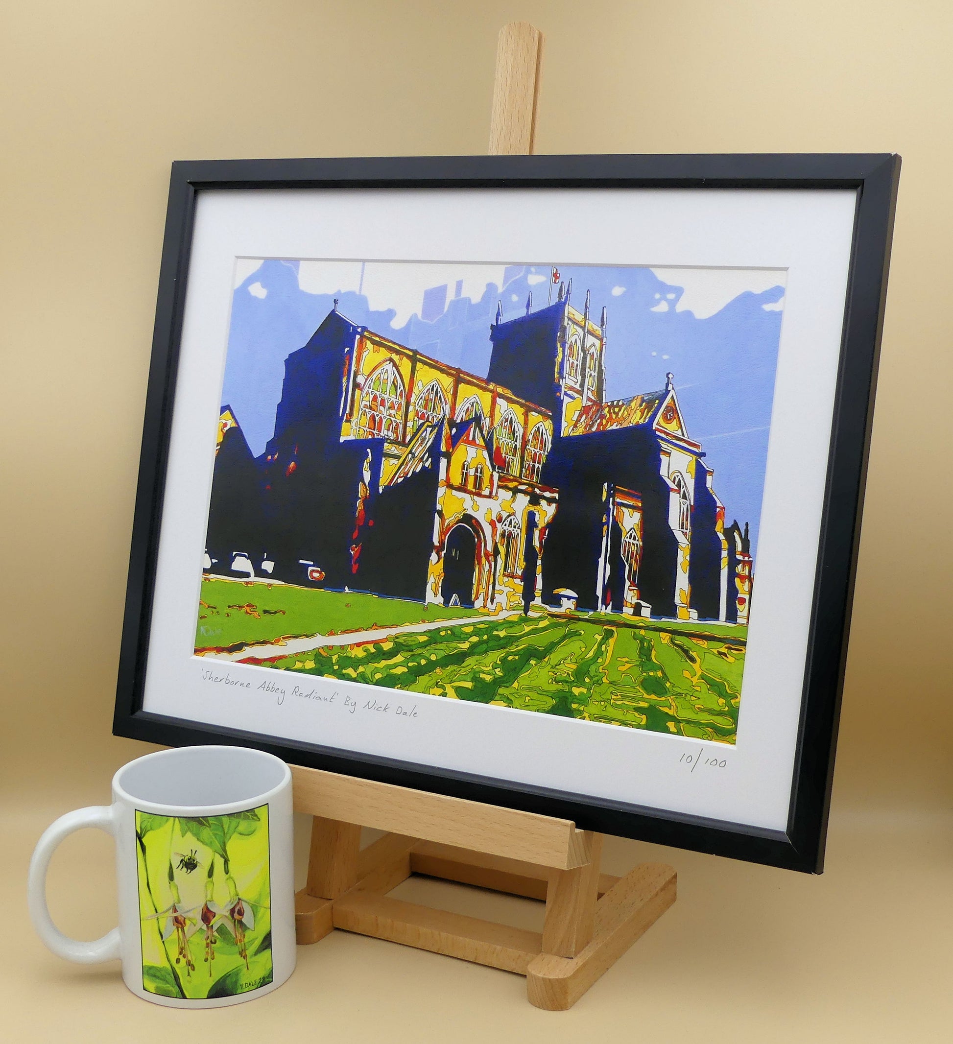 Image of a Medium Art Print featuring a copy of the painting 'Sherborne Abbey Radiant' by Nick Dale. The print depicts Sherborne Abbey in bright primary colours with strong shadows and a brightly lit south-facing aspect. The print is situated on a small easel in a black frame next to a mug.