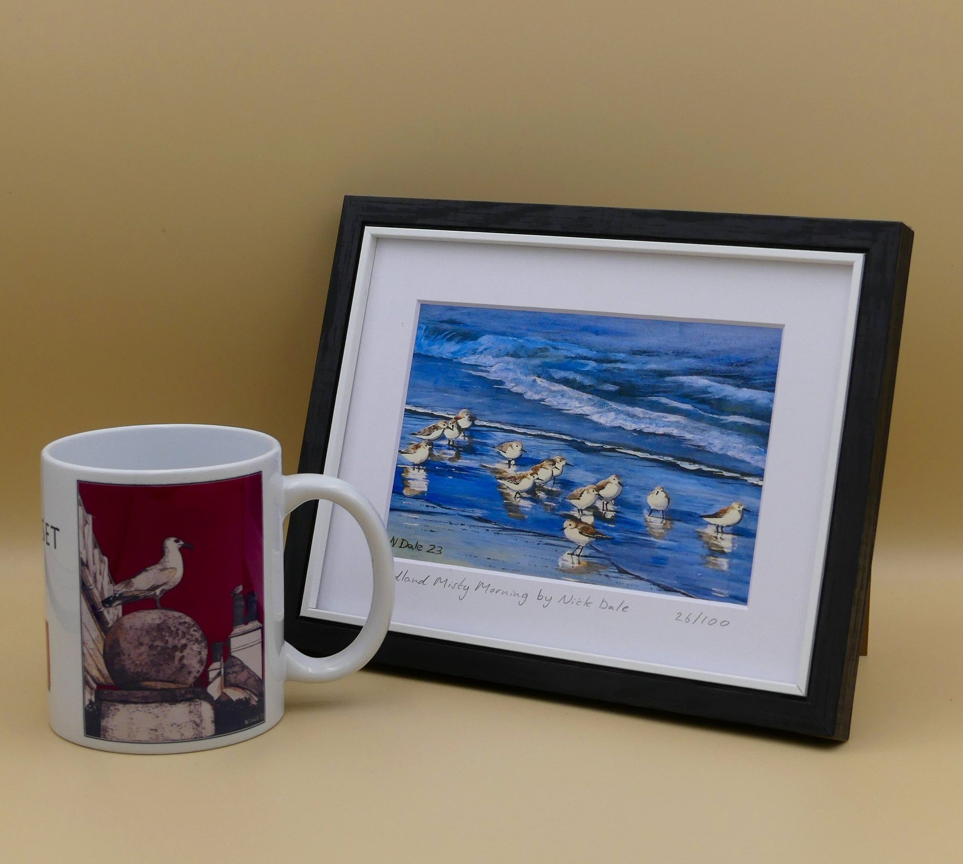 Image of a Small Art Print featuring a copy of the painting 'Studland Misty Morning' by Nick Dale. The print depicts Dunlins on Studland Beach (Dorset) on a crisp and misty morning. The print is situated in a black frame next to a mug.