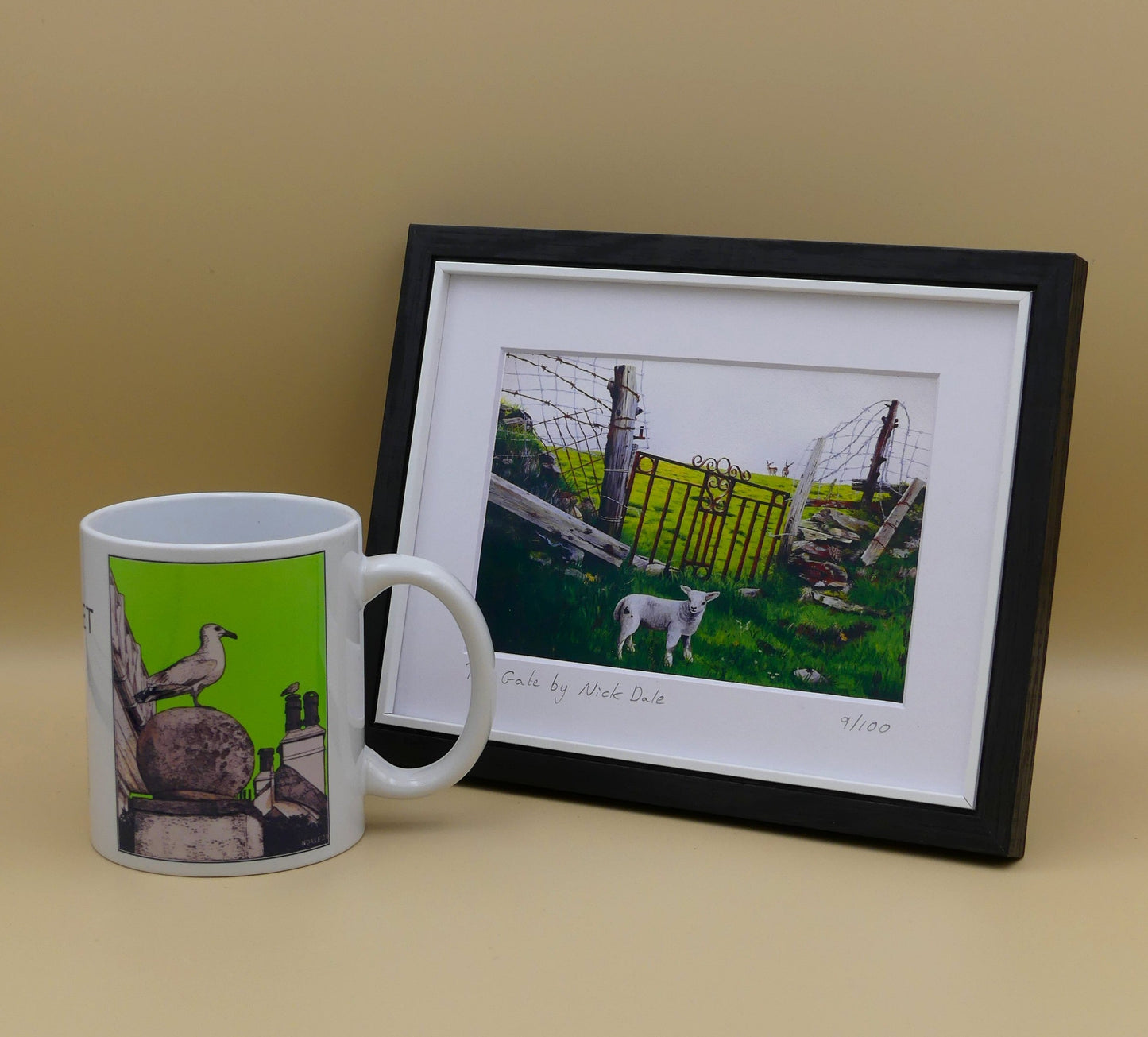Image of a Small Art Print featuring a copy of the painting 'The Gate' by Nick Dale. The print depicts a lamb in front of an ornamental garden gate reused by a farmer somewhere in North Devon, probably to the south of Valley of Rocks, Lynton. The lamb was observed on Lundy Island, Devon.The print is situated in a black frame next to a mug.