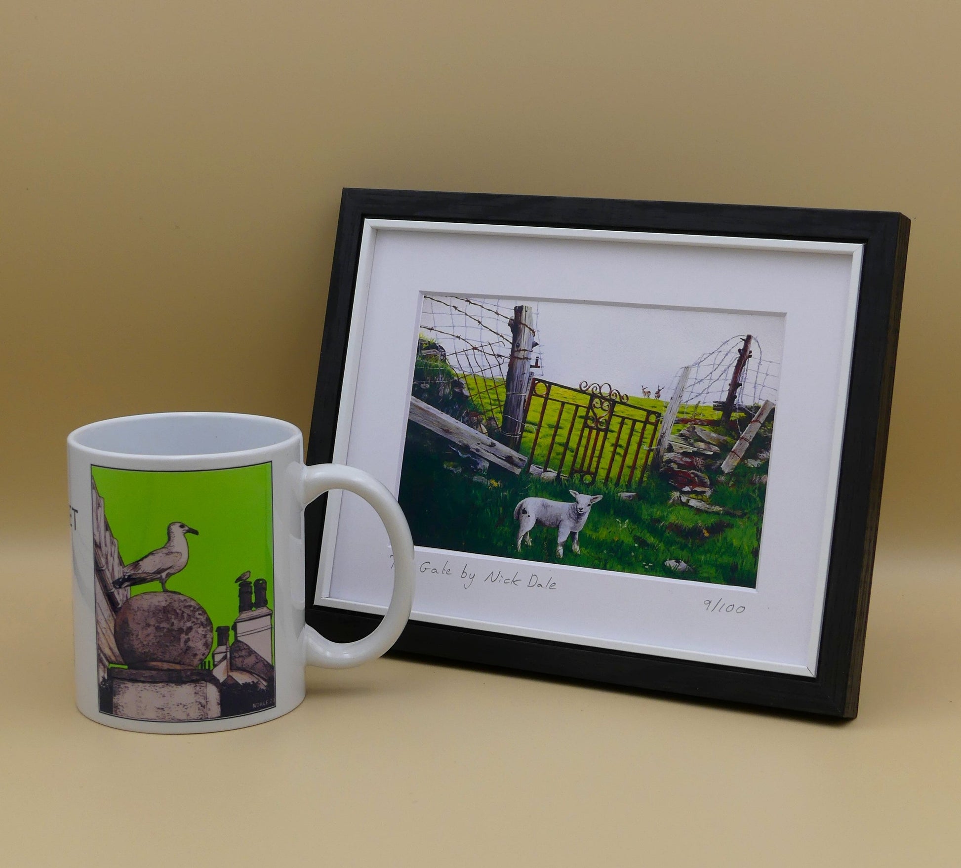 Image of a Small Art Print featuring a copy of the painting 'The Gate' by Nick Dale. The print depicts a lamb in front of an ornamental garden gate reused by a farmer somewhere in North Devon, probably to the south of Valley of Rocks, Lynton. The lamb was observed on Lundy Island, Devon.The print is situated in a black frame next to a mug.