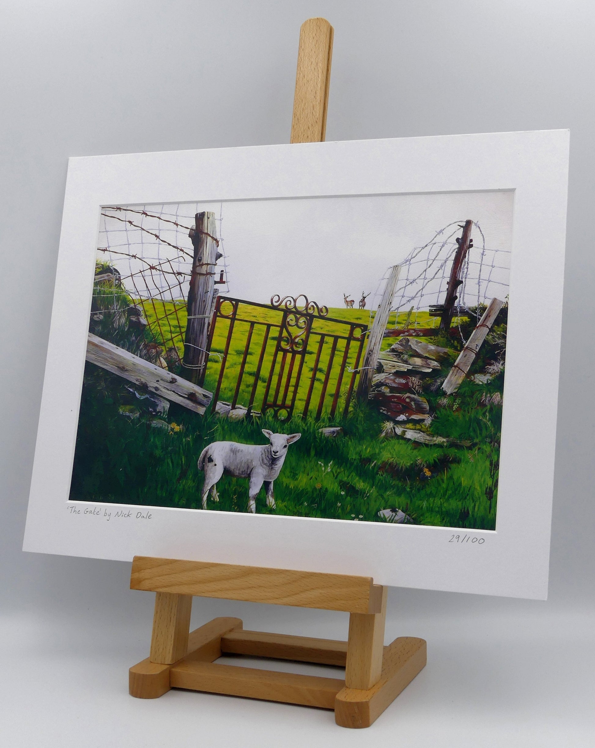 Image of a Medium Art Print featuring a copy of the painting 'The Gate' by Nick Dale. The print depicts a lamb in front of an ornamental garden gate reused by a farmer somewhere in North Devon, probably to the south of Valley of Rocks, Lynton. The lamb was observed on Lundy Island, Devon. The print is situated on a small easel.