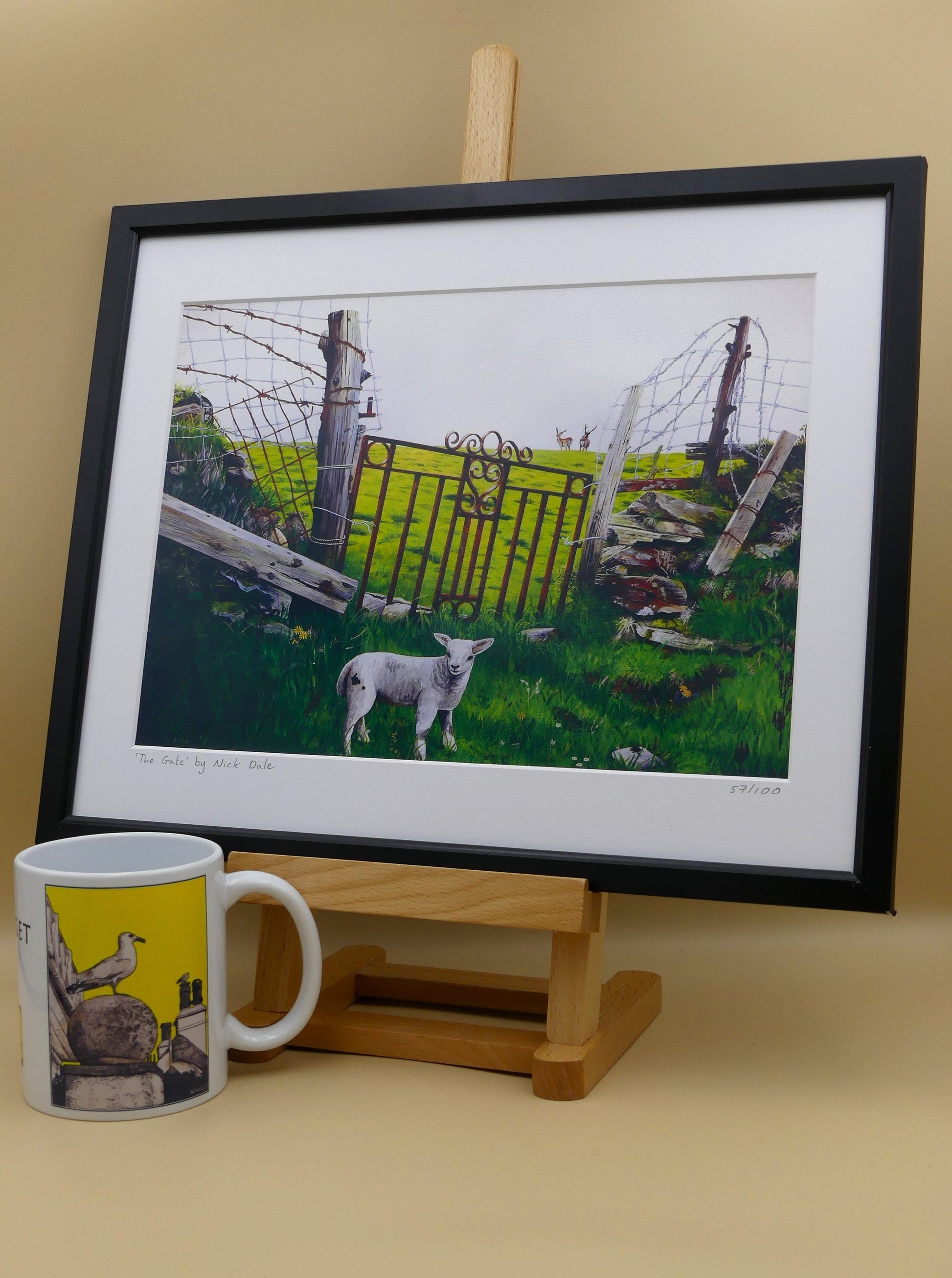 Image of a Medium Art Print featuring a copy of the painting 'The Gate' by Nick Dale. The print depicts a lamb in front of an ornamental garden gate reused by a farmer somewhere in North Devon, probably to the south of Valley of Rocks, Lynton. The lamb was observed on Lundy Island, Devon.The print is situated in a black frame next to a mug.
