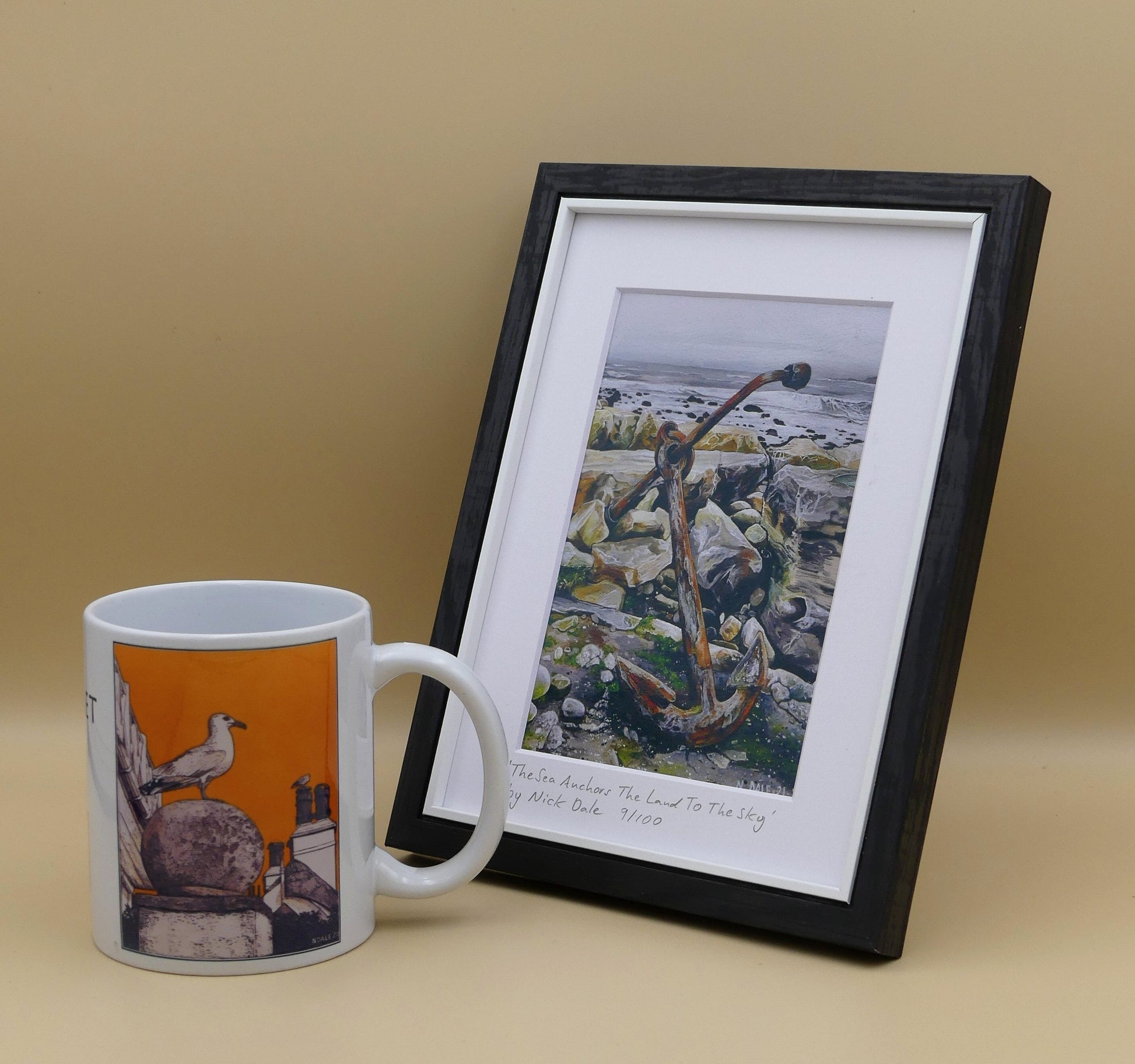 Image of a Small Art Print featuring a copy of the painting 'The Sea Anchors The Land To The Sky' by Nick Dale. The print depicts an old anchor lying at rest under a leaden sky upon the foreshore at Chapman's Pool, near Worth Matravers, Dorset. The print is situated in a black frame next to a mug.