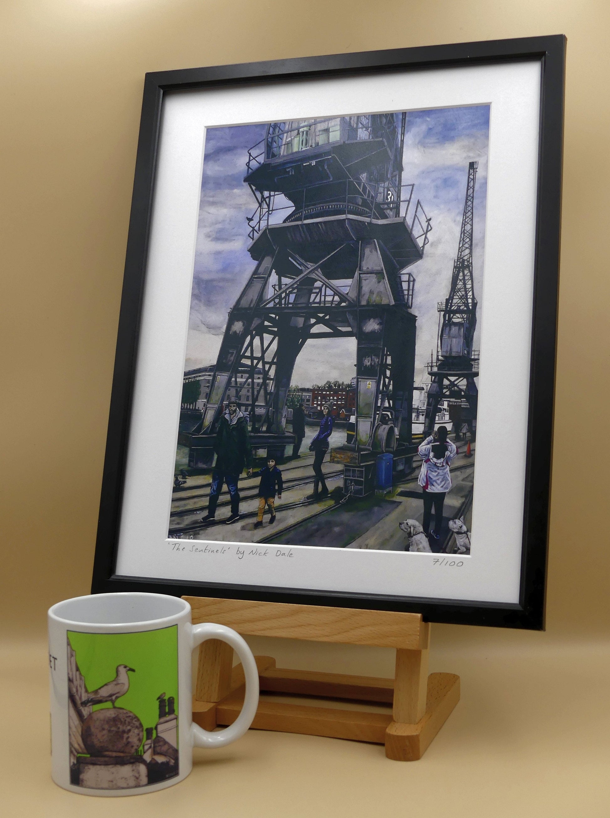 Image of a Medium Art Print featuring a copy of the painting 'The Sentinels' by Nick Dale. The print depicts the iconic tower cranes of Bristol Docks and the classic ship MV Balmoral beyond them. The print is situated in a black frame next to a mug.