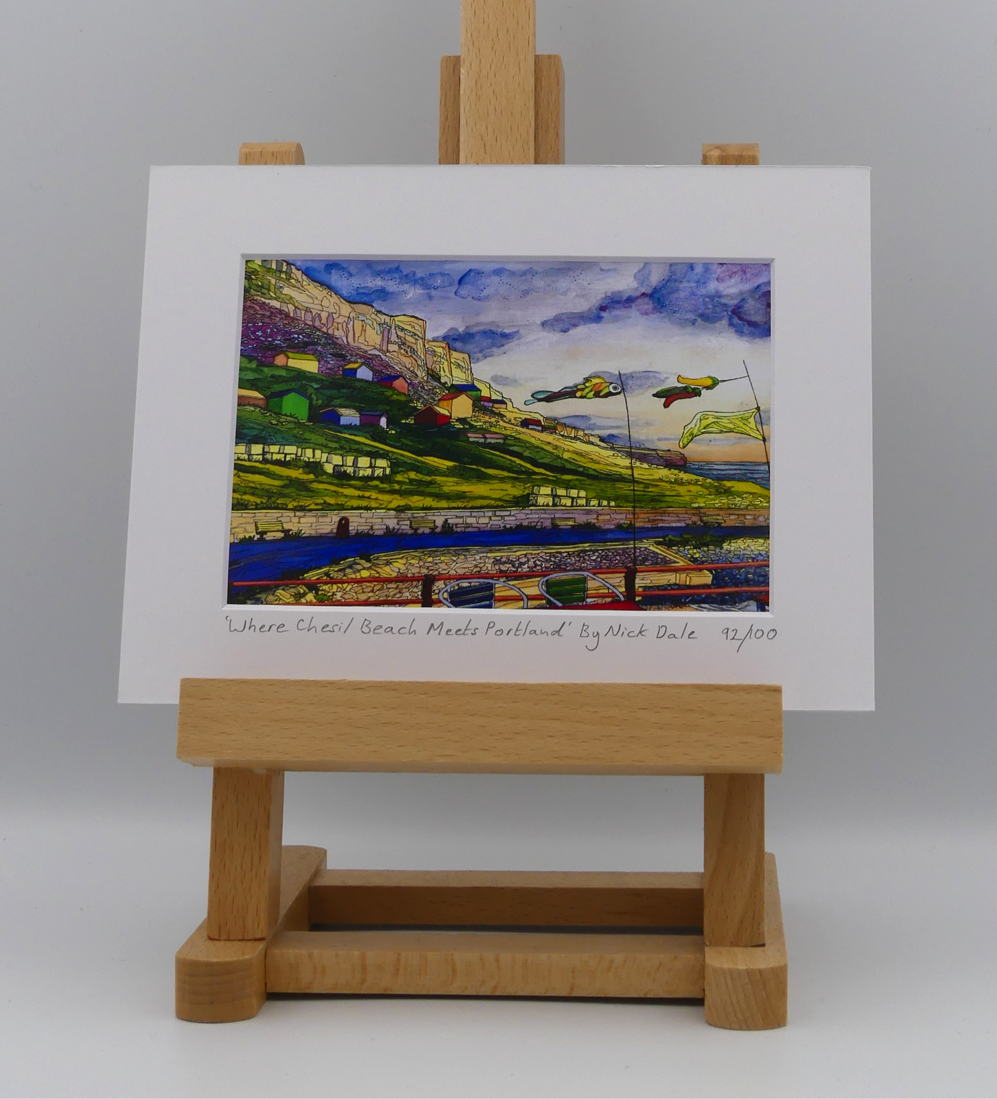 Image of a Small Art Print featuring a copy of the painting 'Where Chesil Beach Meets Portland' by Nick Dale. The print depicts the point where Chesil Beach meets the Isle of Portland, Dorset. In the foreground are the chairs and decorative flags of Quiddles Café Beach Bar. On the slopes below the cliffs of Portland are the many beach huts which overlook the iconic Chesil Cove.  The print is situated on a small easel.