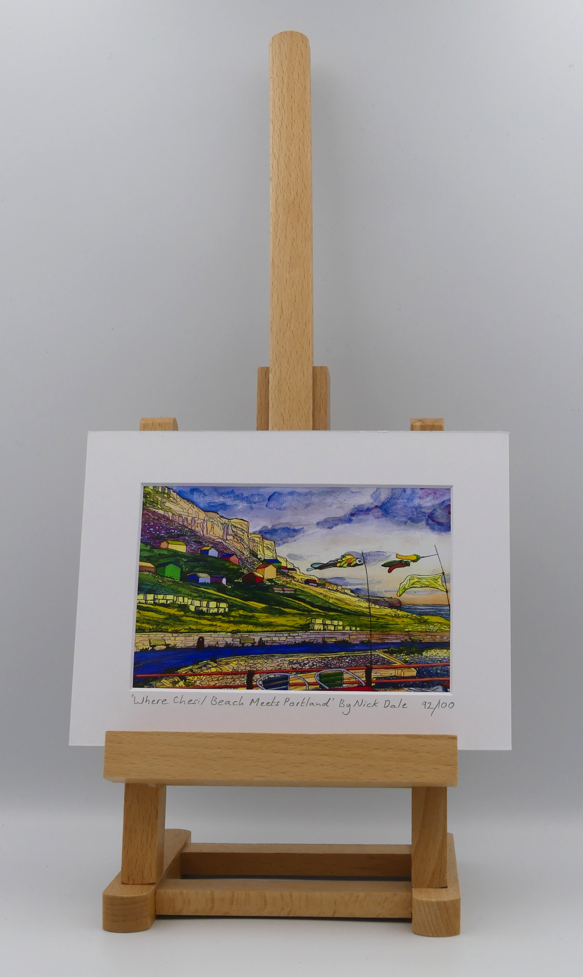 Image of a Small Art Print featuring a copy of the painting 'Where Chesil Beach Meets Portland' by Nick Dale. The print depicts the point where Chesil Beach meets the Isle of Portland, Dorset. In the foreground are the chairs and decorative flags of Quiddles Café Beach Bar. On the slopes below the cliffs of Portland are the many beach huts which overlook the iconic Chesil Cove. The print is situated on a small easel.