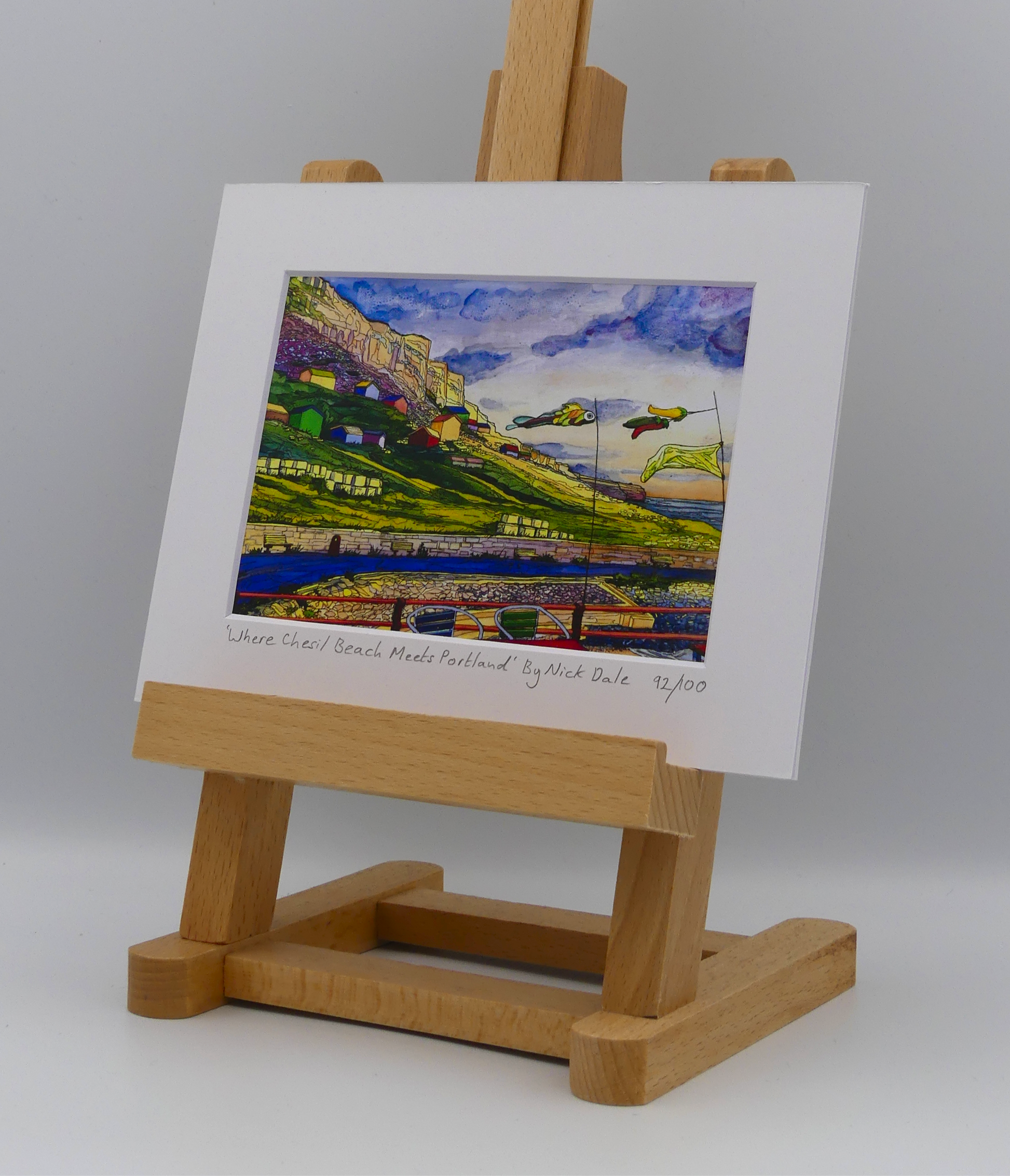 Image of a Small Art Print featuring a copy of the painting 'Where Chesil Beach Meets Portland' by Nick Dale. The print depicts the point where Chesil Beach meets the Isle of Portland, Dorset. In the foreground are the chairs and decorative flags of Quiddles Café Beach Bar. On the slopes below the cliffs of Portland are the many beach huts which overlook the iconic Chesil Cove. The print is situated on a small easel.