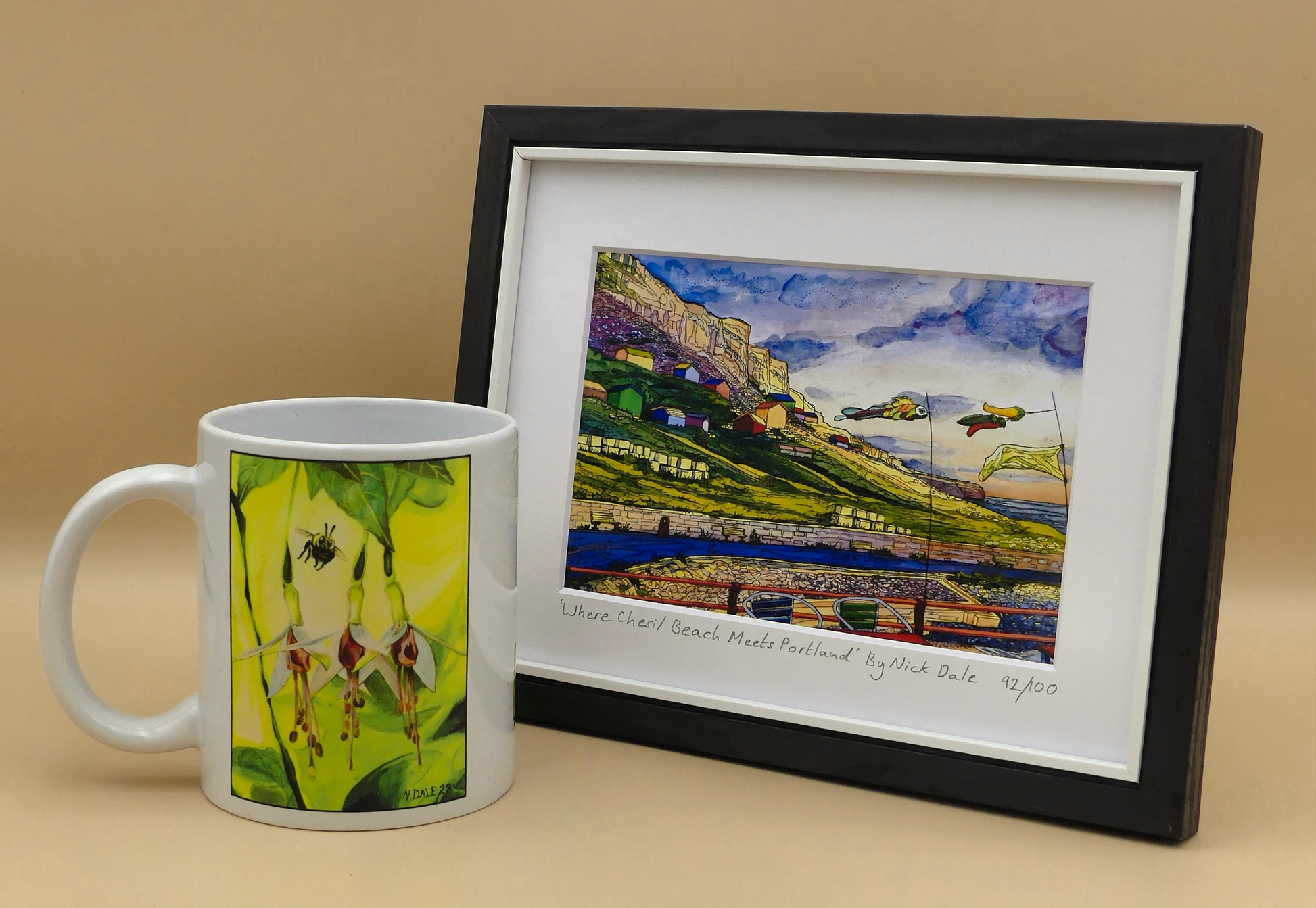 Image of a Small Art Print featuring a copy of the painting 'Where Chesil Beach Meets Portland' by Nick Dale. The print depicts the point where Chesil Beach meets the Isle of Portland, Dorset. In the foreground are the chairs and decorative flags of Quiddles Café Beach Bar. On the slopes below the cliffs of Portland are the many beach huts which overlook the iconic Chesil Cove. The print is situated in a black frame next to a mug.