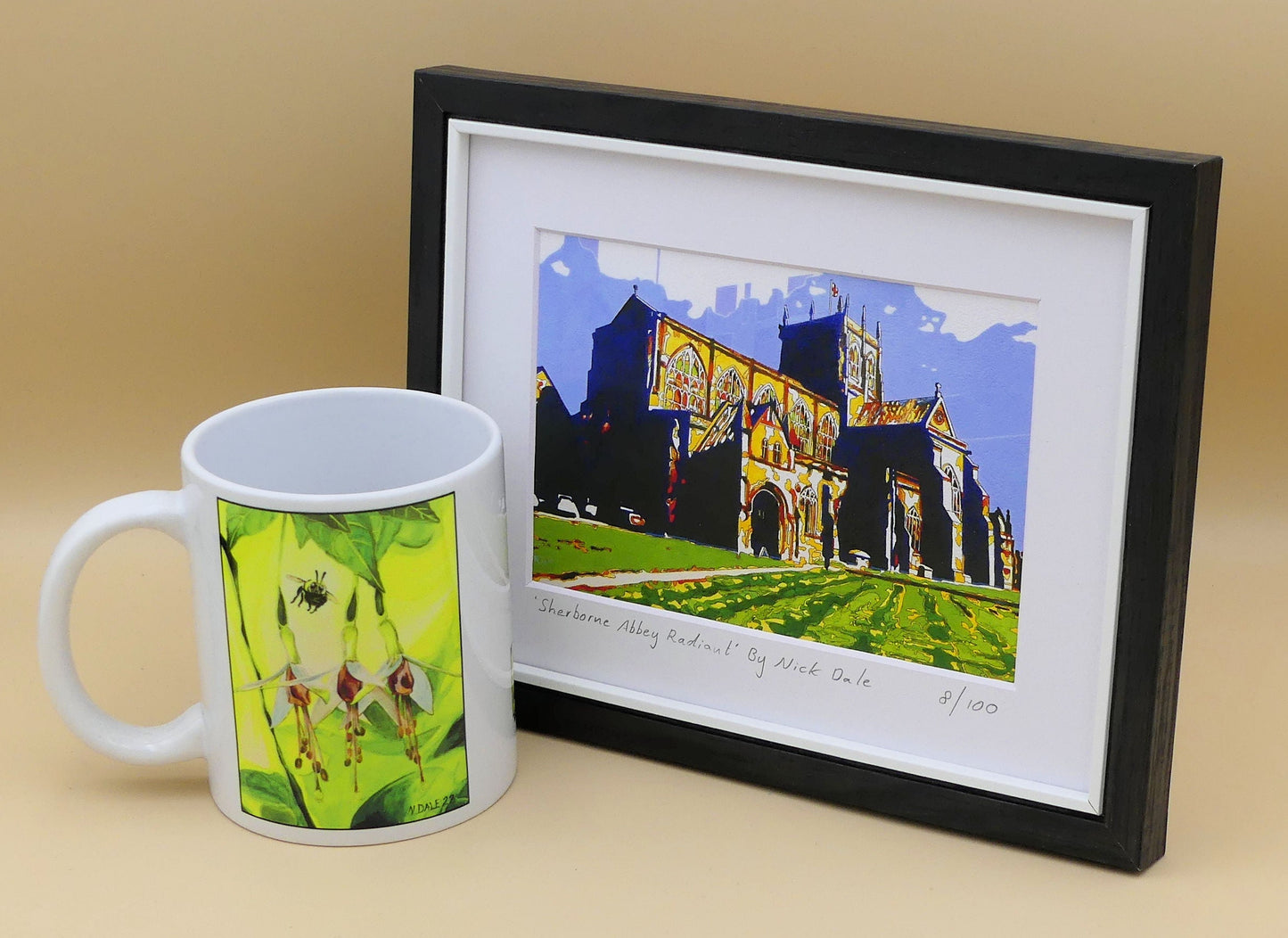 Image of a Small Art Print featuring a copy of the painting 'Sherborne Abbey Radiant' by Nick Dale. The print depicts Sherborne Abbey in bright primary colours with strong shadows and a brightly lit south-facing aspect. The print is situated in a black frame next to a mug.