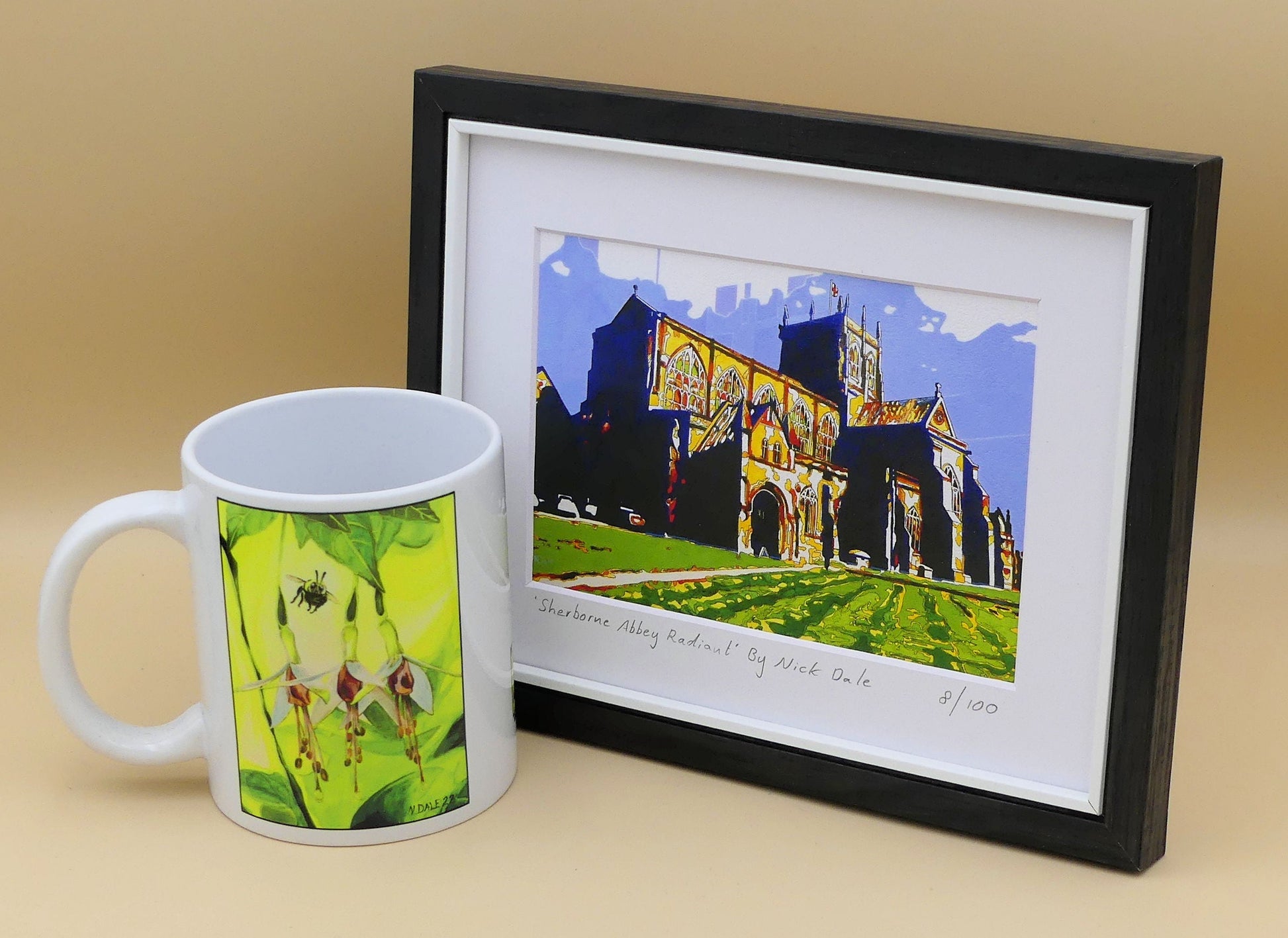 Image of a Small Art Print featuring a copy of the painting 'Sherborne Abbey Radiant' by Nick Dale. The print depicts Sherborne Abbey in bright primary colours with strong shadows and a brightly lit south-facing aspect. The print is situated in a black frame next to a mug.