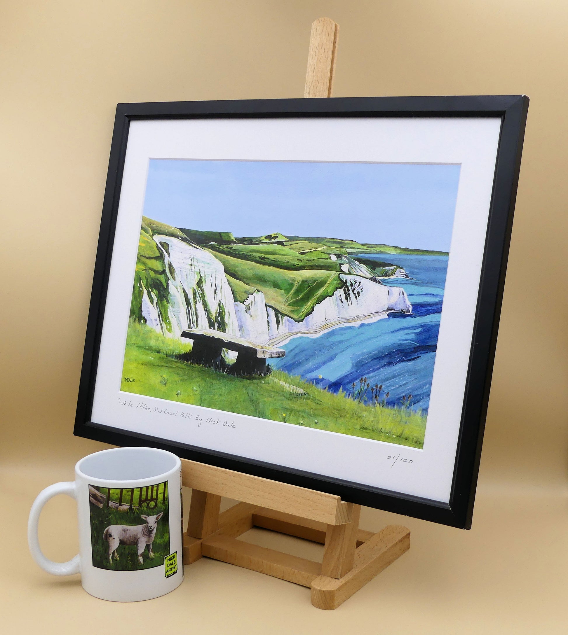 Image of a Medium Art Print featuring a copy of the painting 'White Nothe, SW Coast Path' by Nick Dale. The print depicts a view along the Jurassic Coast from the White Nothe chalk headland east of Ringstead Bay. The view looks east over Bat's Head, Swyre Head and Durdle Door to St Oswald's Bay & beyond. The print is situated on a small easel in a black frame next to a mug.