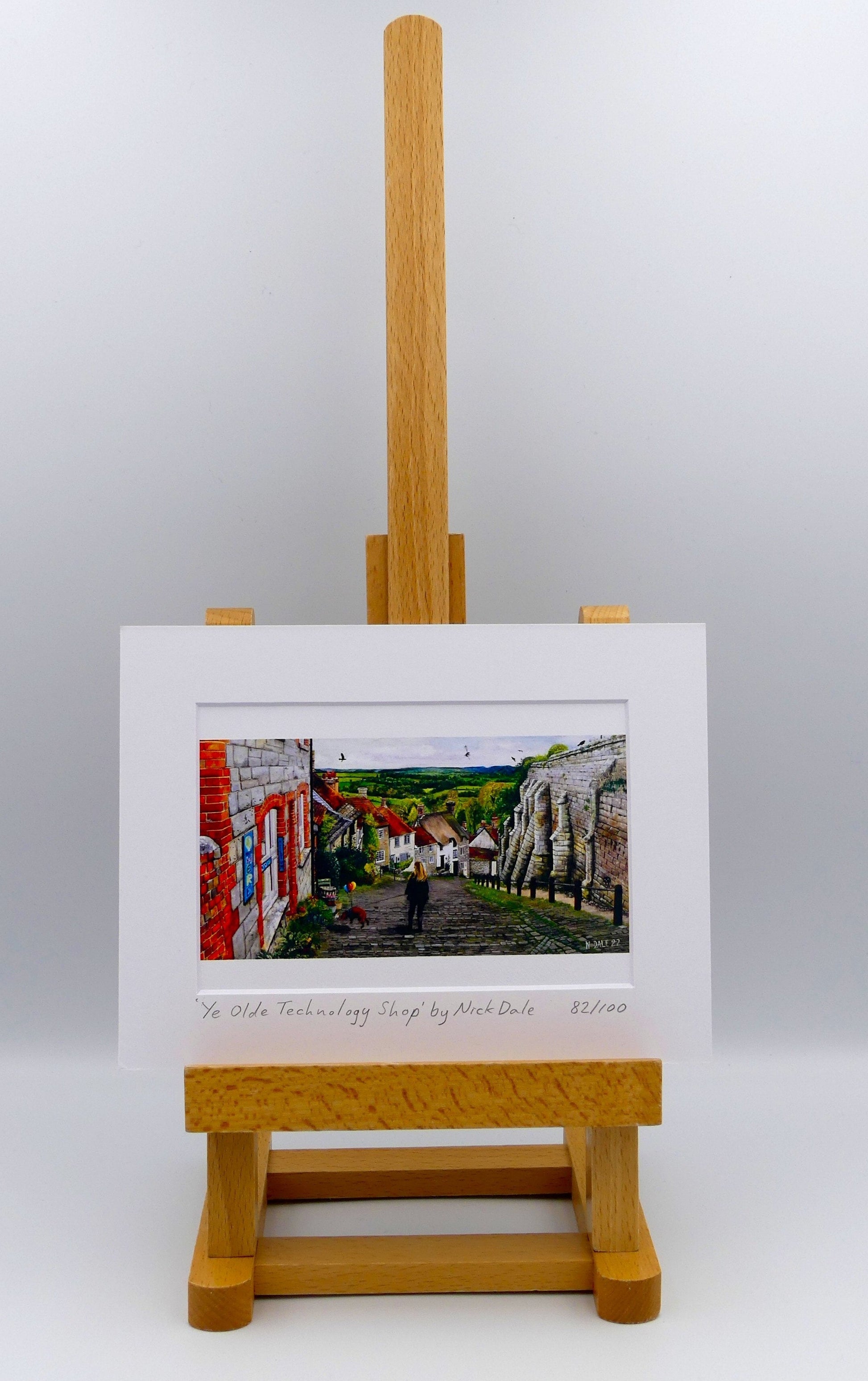 Image of Small Art Print featuring a copy of the painting 'Ye Olde Technology Shop' by Nick Dale. The print depicts the view down Gold Hill in Shaftesbury, Dorset but in a slightly surreal form as the scene depicts a shop selling helium used to enable dogs to float. The dog is trying to get to a dropped ice-cream cone but is unable to reach as it floats above it. The print is situated on a small easel.