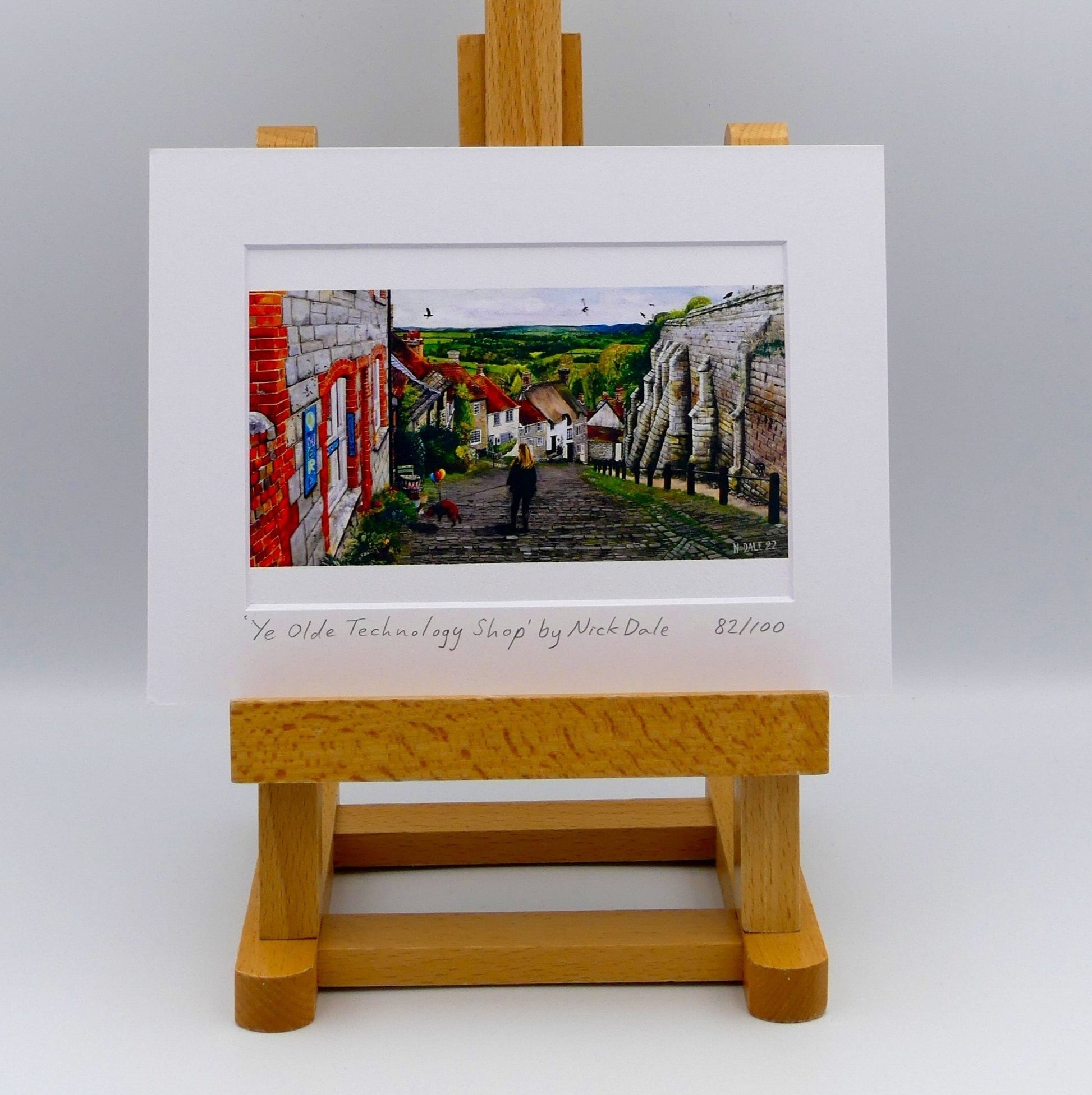 Image of Small Art Print featuring a copy of the painting 'Ye Olde Technology Shop' by Nick Dale. The print depicts the view down Gold Hill in Shaftesbury, Dorset but in a slightly surreal form as the scene depicts a shop selling helium used to enable dogs to float. The dog is trying to get to a dropped ice-cream cone but is unable to reach as it floats above it. The print is situated on a small easel.