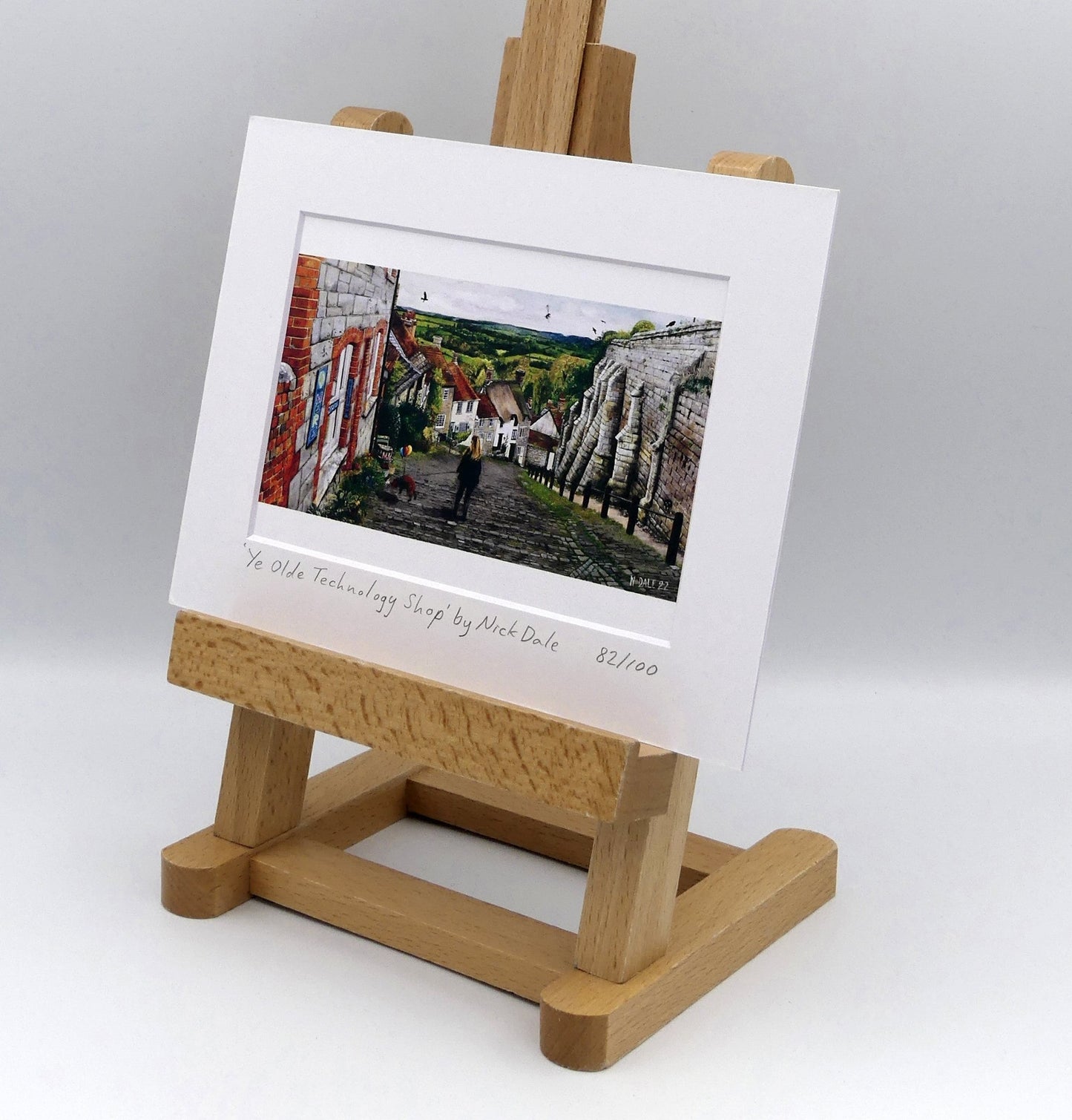 Image of Small Art Print featuring a copy of the painting 'Ye Olde Technology Shop' by Nick Dale. The print depicts the view down Gold Hill in Shaftesbury, Dorset but in a slightly surreal form as the scene depicts a shop selling helium used to enable dogs to float. The dog is trying to get to a dropped ice-cream cone but is unable to reach as it floats above it. The print is situated on a small easel.