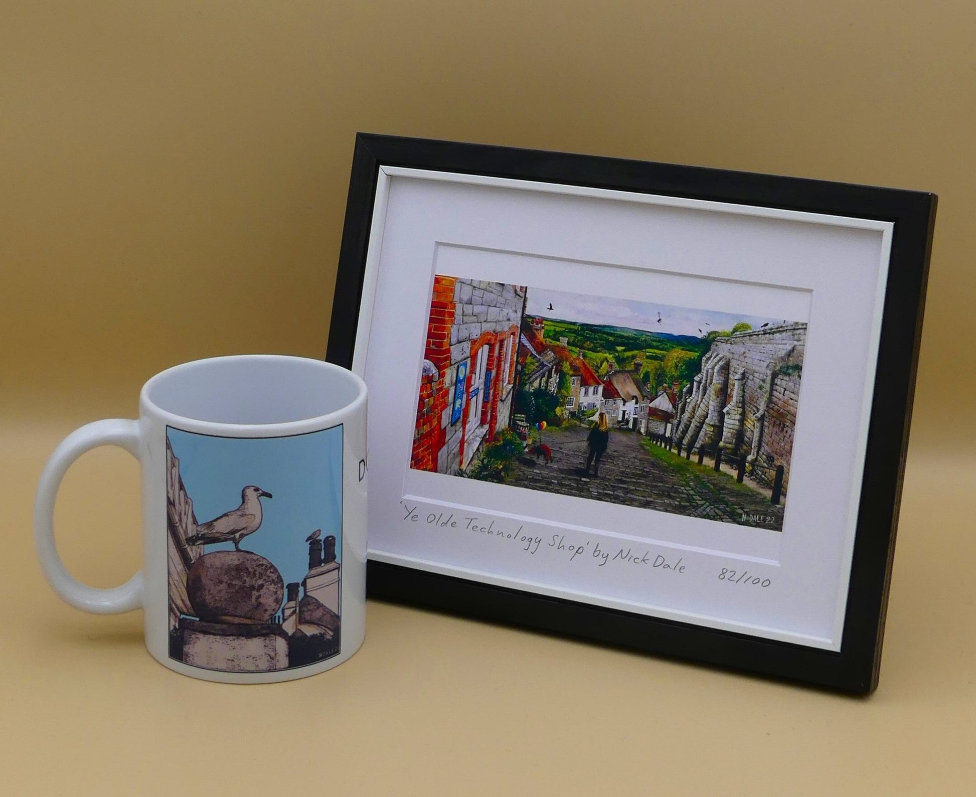 Image of Small Art Print featuring a copy of the painting 'Ye Olde Technology Shop' by Nick Dale. The print depicts the view down Gold Hill in Shaftesbury, Dorset but in a slightly surreal form as the scene depicts a shop selling helium used to enable dogs to float. The dog is trying to get to a dropped ice-cream cone but is unable to reach as it floats above it. The print is situated in a black frame next to a mug.