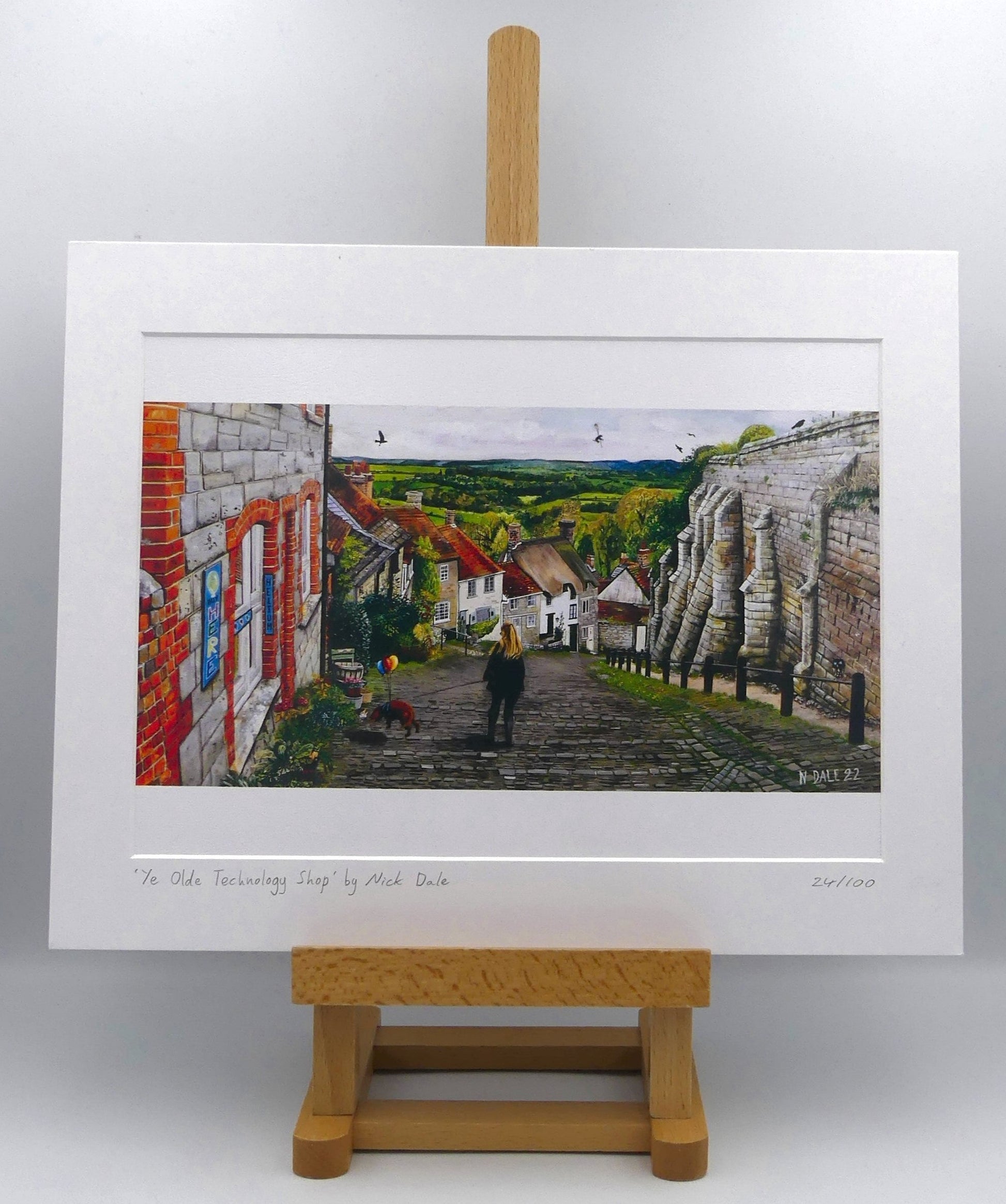 Image of Medium Art Print featuring a copy of the painting 'Ye Olde Technology Shop' by Nick Dale. The print depicts the view down Gold Hill in Shaftesbury, Dorset but in a slightly surreal form as the scene depicts a shop selling helium used to enable dogs to float. The dog is trying to get to a dropped ice-cream cone but is unable to reach as it floats above it. The print is situated on a small easel.