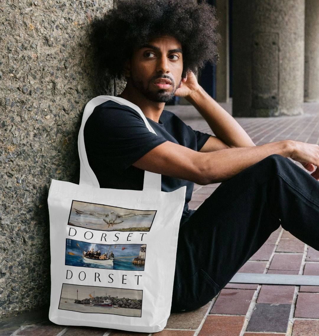 Image of male model holding White Tote Bag featuring 3 Scenes Of Dorset. The scenes are derived from 3 original paintings by Nick Dale  and are separated from each other vertically by the word 'Dorset'. 