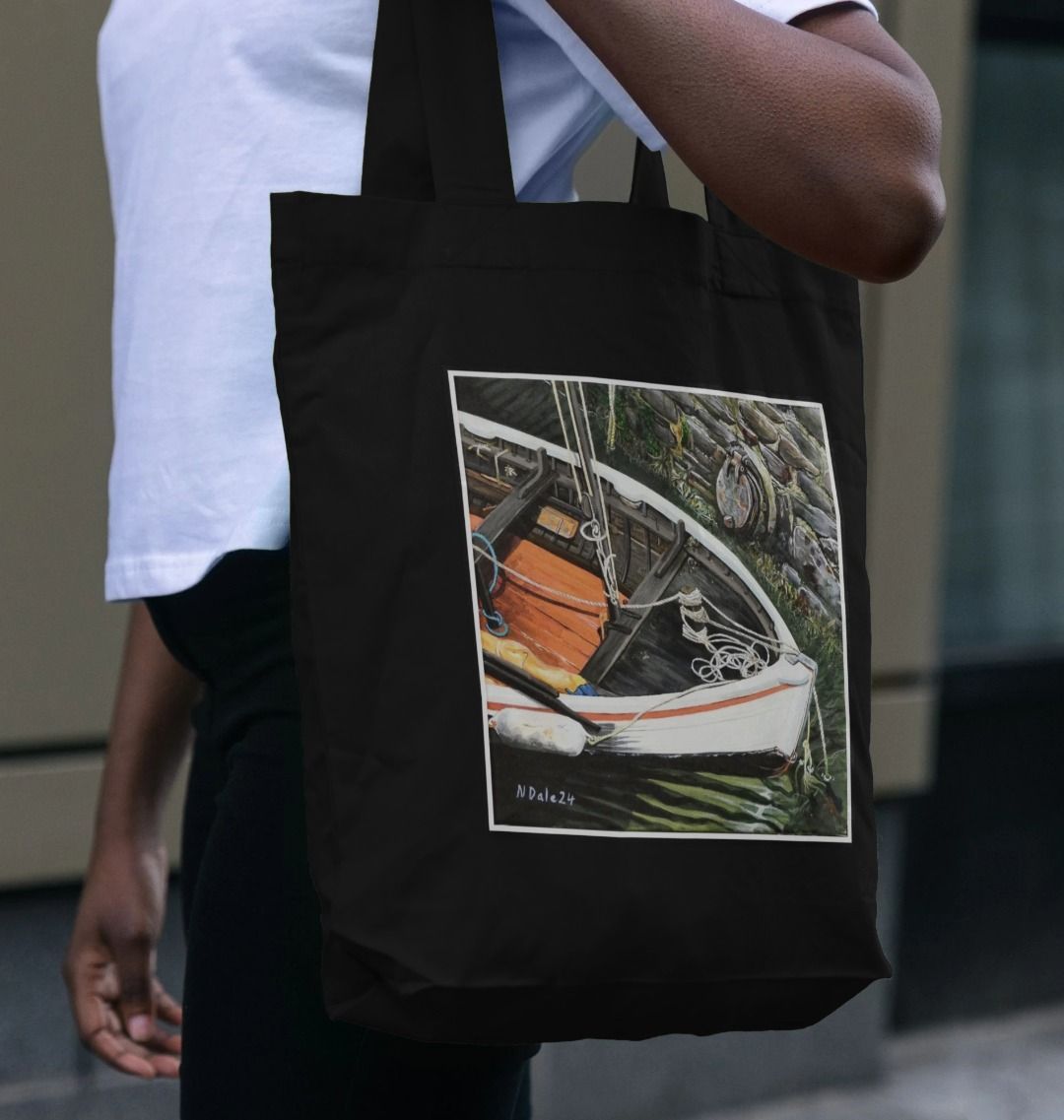Image of a female model carrying a Black Tote Bag featuring a copy of the painting 'The Outlet' by Nick Dale. The bag depicts a view down into the harbour at Lynmouth in North Devon showing a small sailing boat at rest against the harbour wall next to a water outfall pipe.