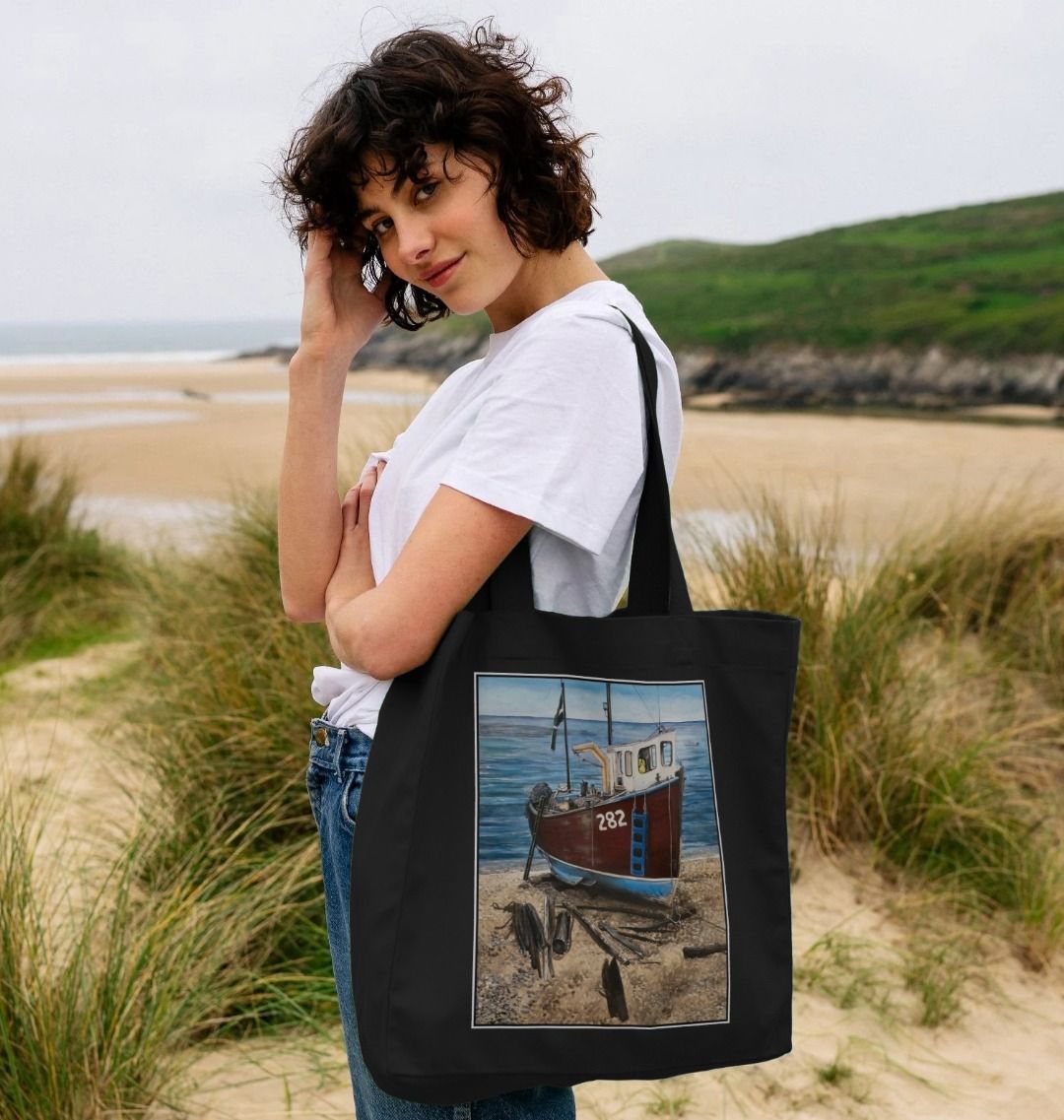 Image of female model carrying a Black Shopper Tote Bag featuring a copy of the painting 'Relaxing Beer' by Nick Dale. The bag depicts a fishing boat beached at Beer, Devon. The wood is laid under the boats to aid in their winching up the shingle beach.