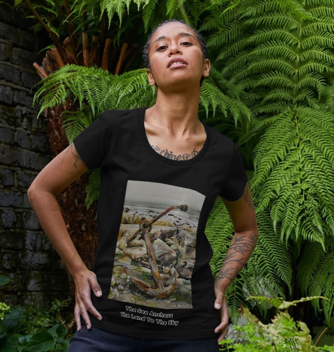 Image of a female model wearing a Black Women's Scoop Neck T-shirt featuring a copy of the painting 'The Sea Anchors The Land To The Sky' by Nick Dale, with the words 'The Sea Anchors The Land To The Sky' written beneath it. The T-shirt depicts an old anchor lying at rest under a leaden sky upon the foreshore at Chapman's Pool, near Worth Matravers, Dorset.