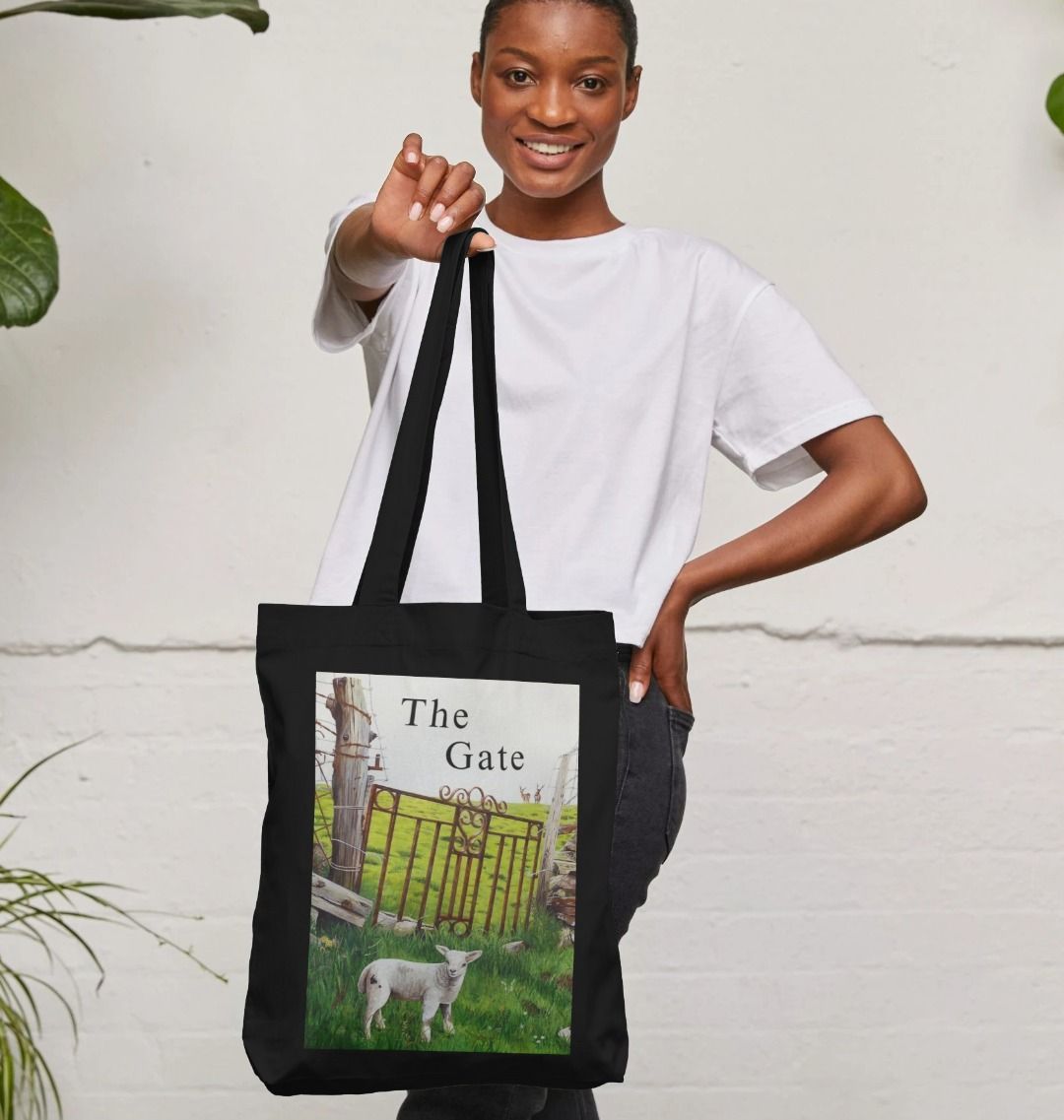Image of a female model holding a Black Tote Bag featuring a copy of a detail from the painting 'The Gate' by Nick Dale, with the words 'the Gate' written above the gate. The bag depicts a lamb in front of an ornamental garden gate reused by a farmer somewhere in North Devon, probably to the south of Valley of Rocks, Lynton. The lamb was observed on Lundy Island, Devon.