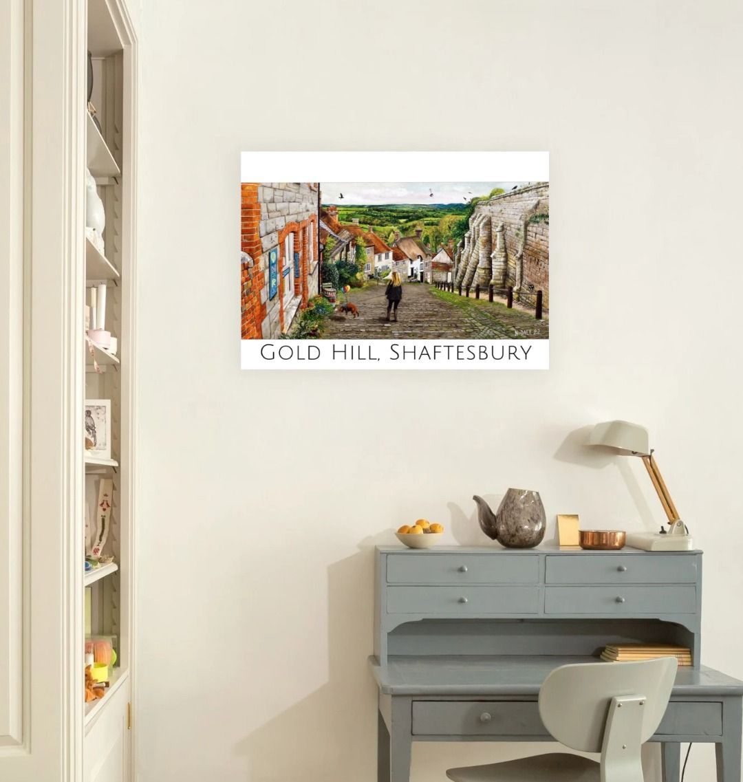 Image of a Large Art Print (A3 size) featuring a copy of the painting 'Ye Olde Technology Shop' by Nick Dale, with the words 'Gold Hill, Shaftesbury' written beneath it; as displayed on a wall above a desk. The print depicts the view down Gold Hill in Shaftesbury, Dorset but in a slightly surreal form as the scene depicts a shop selling helium used to enable dogs to float. The dog is trying to get to a dropped ice-cream cone but is unable to reach as it floats above it.