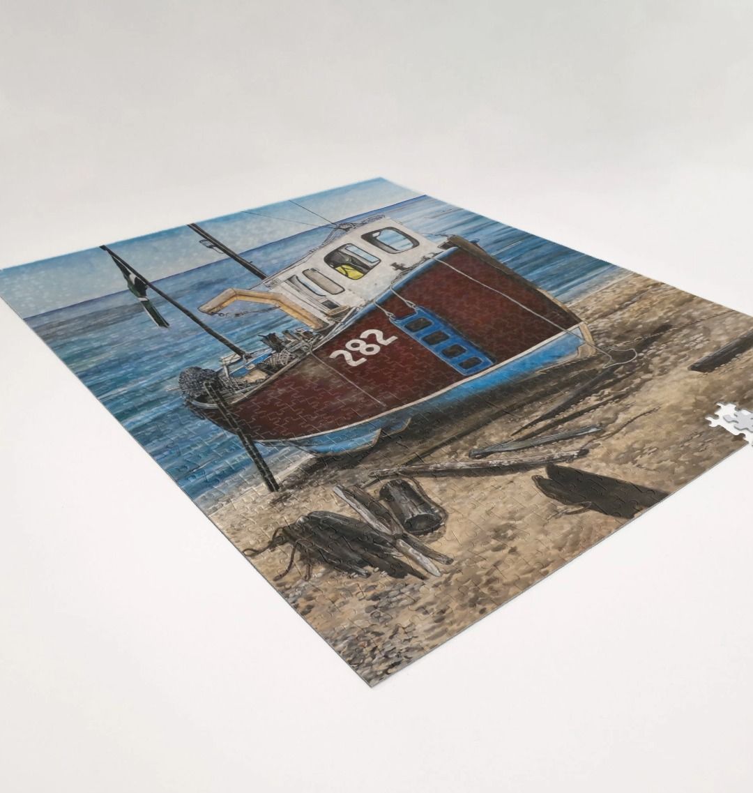 Image of a 1000 Piece Jigsaw Puzzle featuring a copy of the painting 'Relaxing Beer' by Nick Dale. As displayed partially completed on a white surface. The jigsaw depicts a fishing boat beached at Beer, Devon. The wood is laid under the boats to aid in their winching up the shingle beach.