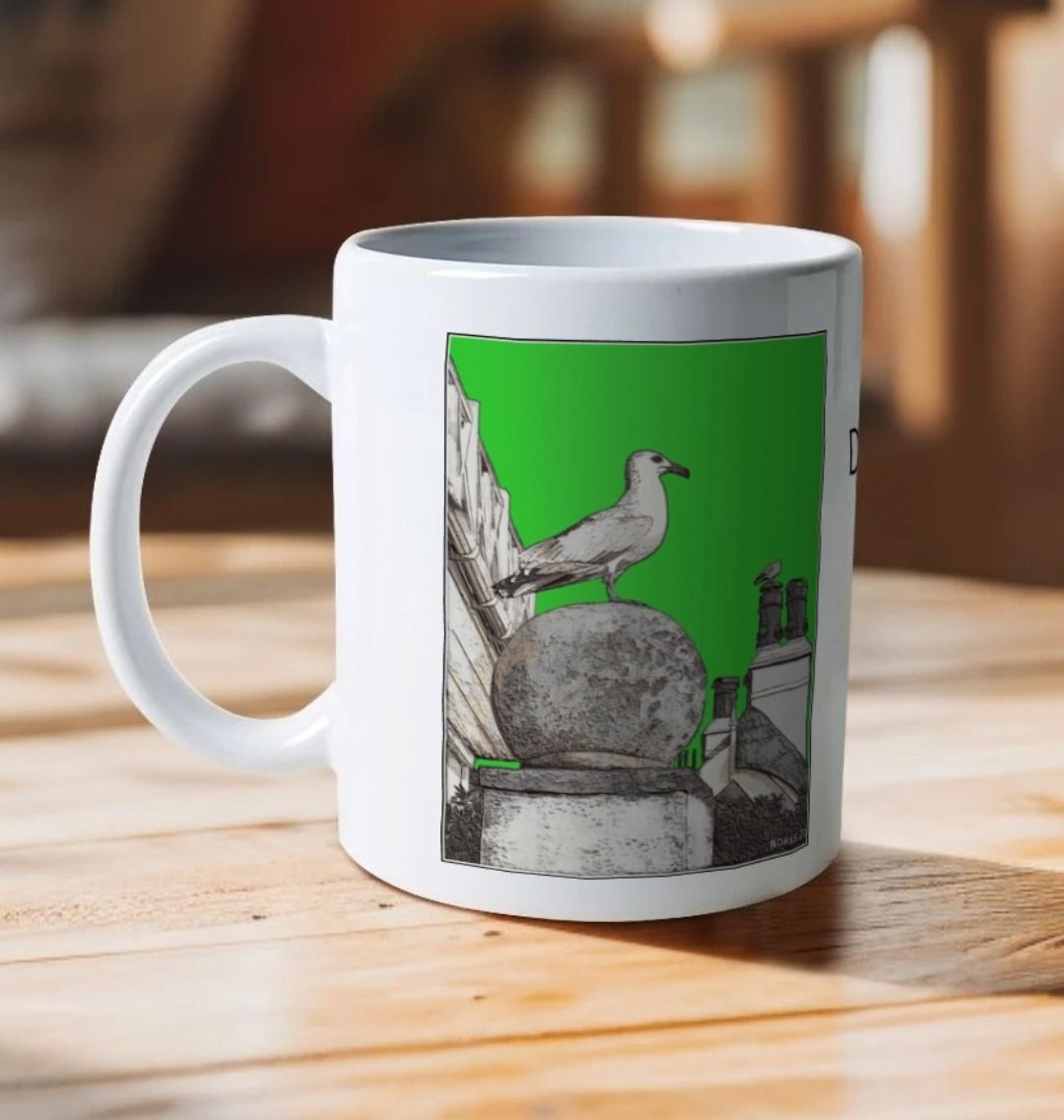 Image of a White Mug on a wooden table featuring a modified copy of the painting 'A View Of The Sea At Lyme Regis' by Nick Dale - this being the 'Green' version. The handle is orientated to the left. The mug depicts a gull and buildings along Marine Parade, Lyme Regis.