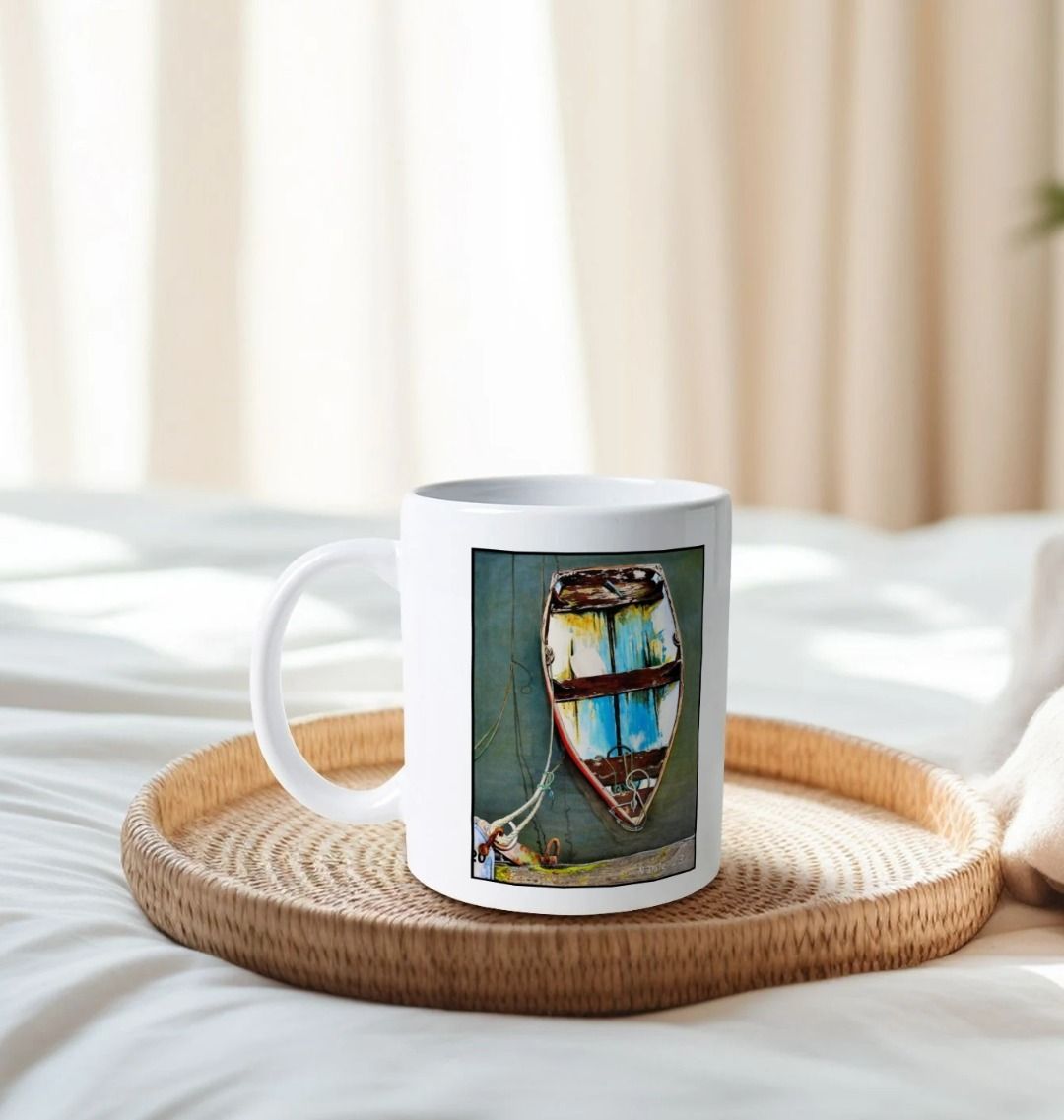 Image of a White Mug on a tray on a bed featuring a copy of the painting 'Good Intentions at Lyme Regis' by Nick Dale. The handle is orientated to the left. The mug depicts a small wooden rowing boat tied up in Lyme Regis harbour, Dorset.