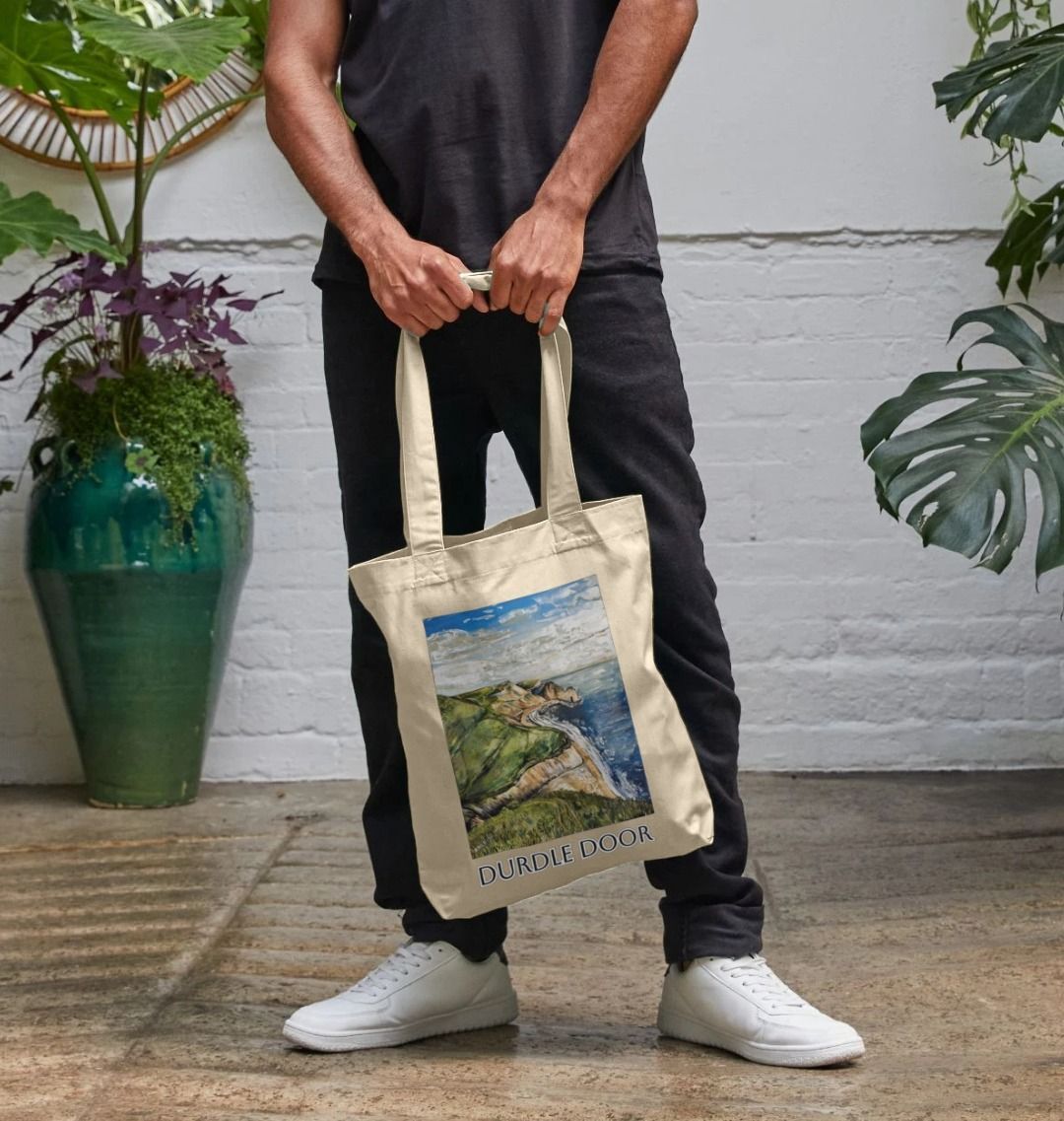 Image of male model carrying a Natural Tote Bag featuring a copy of the painting 'Durdle Door From The West' by Nick Dale with the words 'Durdle Door' written below it. The bag depicts a view along the Jurassic Coast to the famous Dorset limestone arch landmark, as seen from the coastal path to the west.