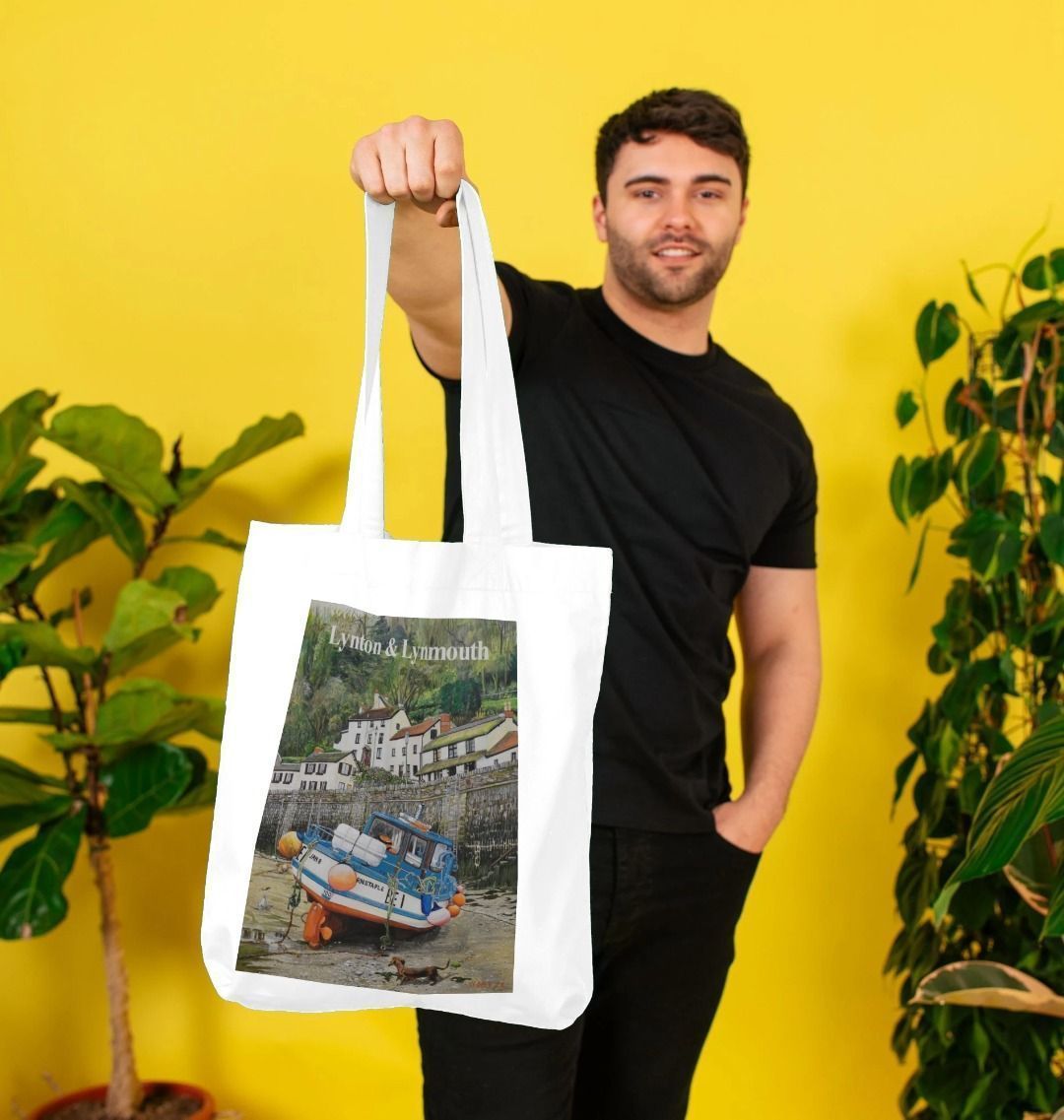 Image of a male model holding a White Tote Bag featuring a copy of the painting 'Lynmouth At Lowest Tide' by Nick Dale, with the words 'Lynton & Lynmouth' written across the top. The bag depicts an unusual perspective on the picturesque harbour of Lynmouth (Devon), as seen from the floor of the harbour at low tide. Behind the fishing boat can be seen the characterful harbourside buildings.
