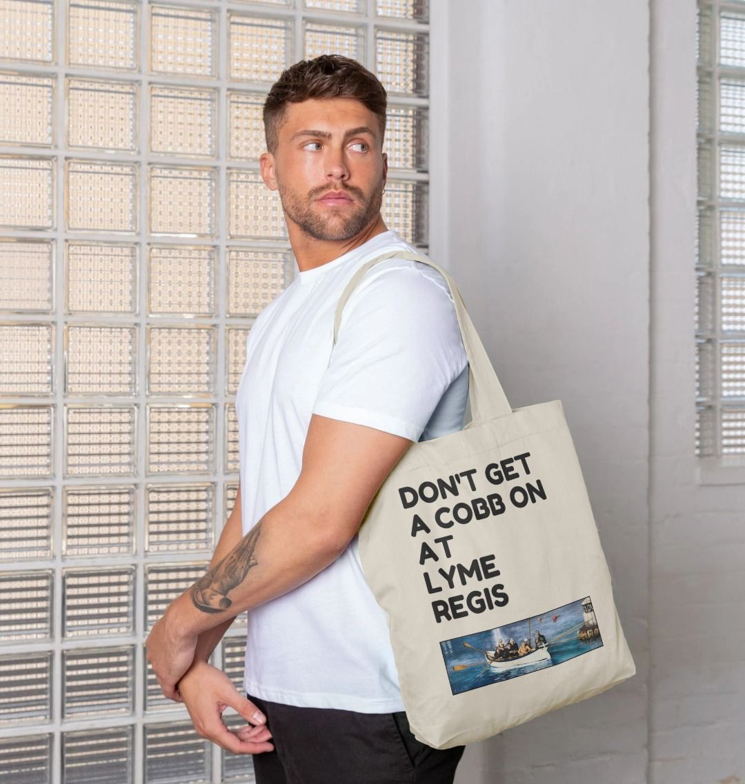Image of a male model carrying a Natural Tote Bag featuring a copy of the painting 'The Return to Base at Lyme Regis' by Nick Dale with the slogan 'Don't Get A Cobb On At Lyme Regis' written above it. The bag depicts the crew of a gig rowing boat returning to Lyme Regis Harbour to take a well-earned rest.