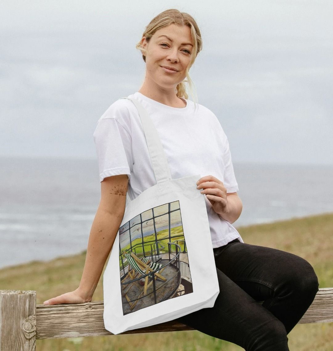 Image of a female model carrying a White Tote Bag featuring a copy of the painting 'Let's Be Together Alone' by Nick Dale. The bag depicts the view from the lantern room of the Old Light lighthouse on Beacon Hill, on the island of Lundy, Devon.