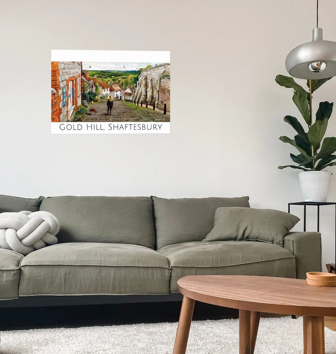 Image of a Large Art Print (A3 size) featuring a copy of the painting 'Ye Olde Technology Shop' by Nick Dale, with the words 'Gold Hill, Shaftesbury' written beneath it; as displayed on a wall above a sofa. The print depicts the view down Gold Hill in Shaftesbury, Dorset but in a slightly surreal form as the scene depicts a shop selling helium used to enable dogs to float. The dog is trying to get to a dropped ice-cream cone but is unable to reach as it floats above it.