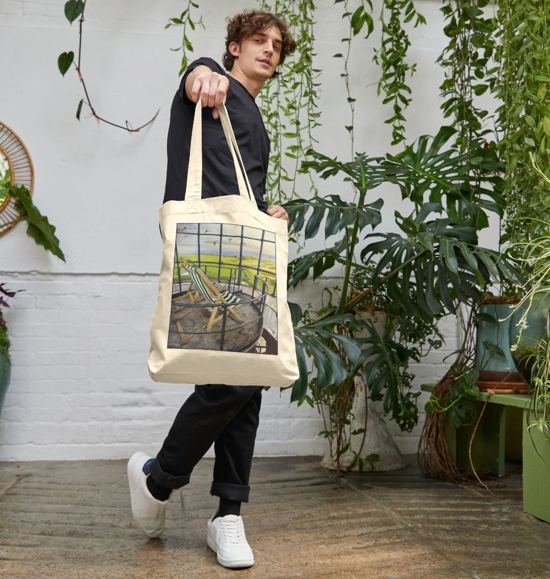 Image of a male model holding a Natural Tote Bag featuring a copy of the painting 'Let's Be Together Alone' by Nick Dale. The bag depicts the view from the lantern room of the Old Light lighthouse on Beacon Hill, on the island of Lundy, Devon.