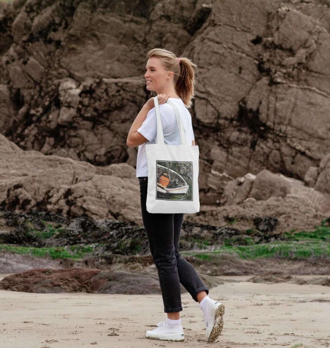 Image of a female model carrying a White Tote Bag featuring a copy of the painting 'The Outlet' by Nick Dale. The bag depicts a view down into the harbour at Lynmouth in North Devon showing a small sailing boat at rest against the harbour wall next to a water outfall pipe.