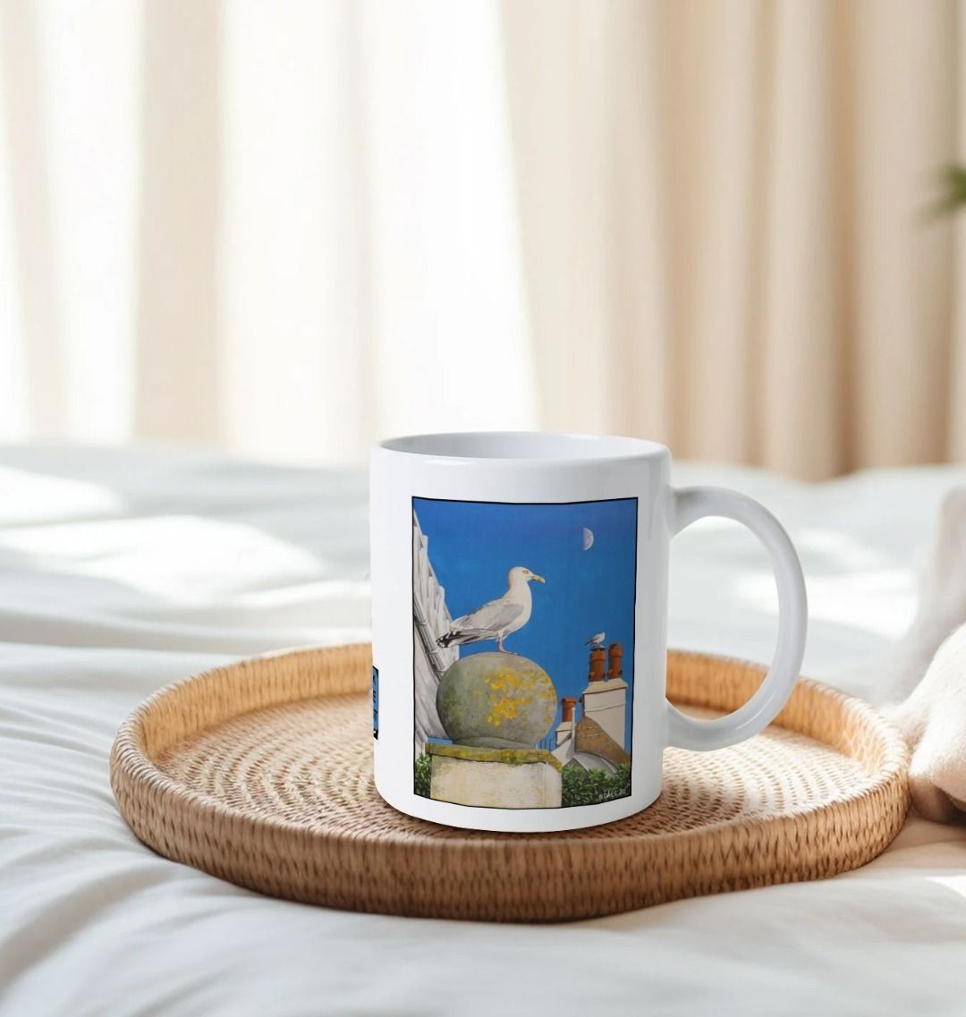 Image of a White Mug on a tray on a bed featuring a copy of the painting 'A View Of The Sea At Lyme Regis' by Nick Dale. The handle is orientated to the right. The mug depicts a gull and buildings along Marine Parade, Lyme Regis.