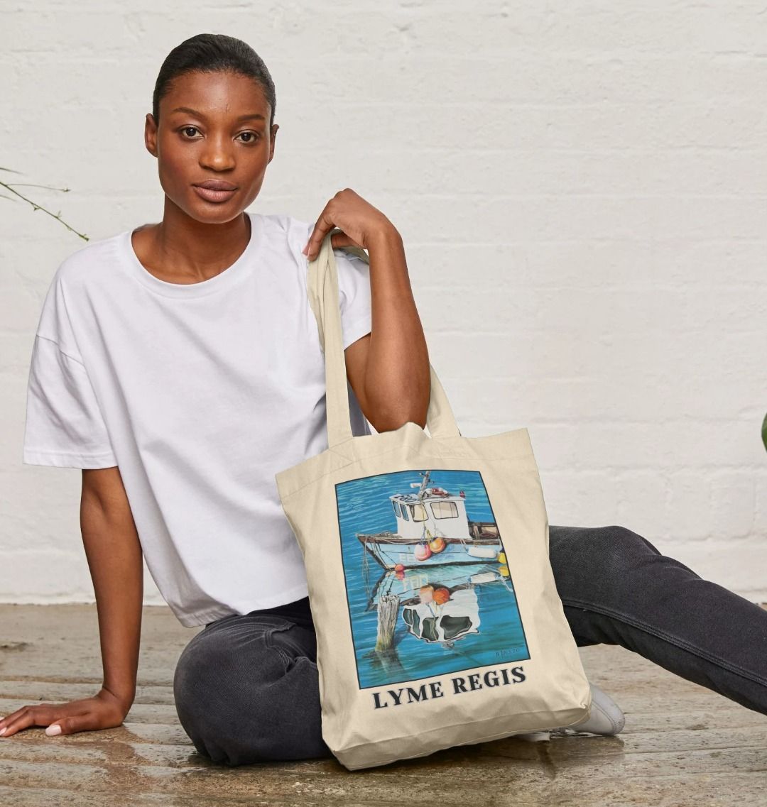 Image of a female model holding a Natural Tote Bag featuring a copy of the painting 'God's Own 99 Flake Pierces The Veil' by Nick Dale, with the words 'Lyme Regis' written beneath it. The bag depicts a fishing boat at rest within the harbour of Lyme Regis, Dorset. The title is based on the similarity of the wooden pile to a chocolate flake as inserted into an ice cream cone. This seemed appropriate given the draw of the town to tourists.